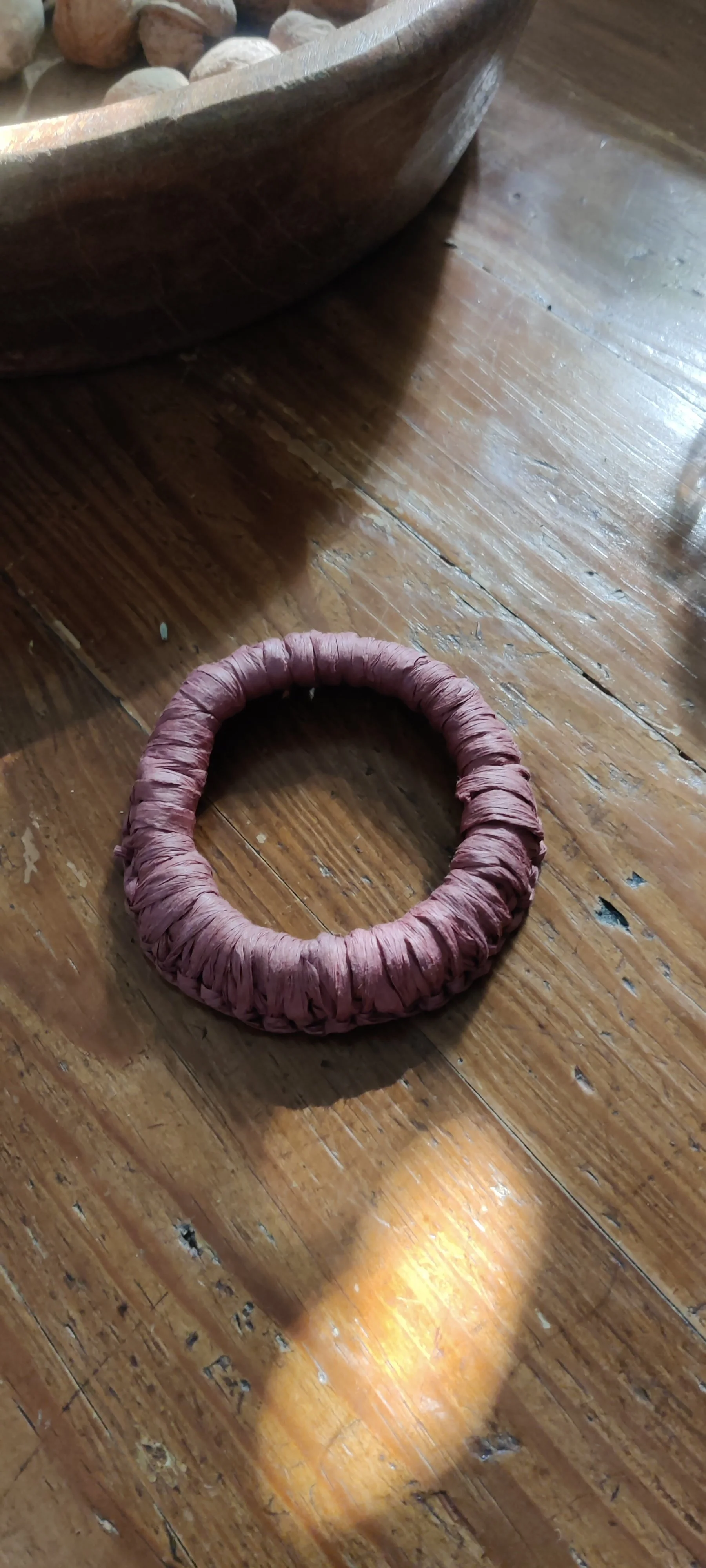 Pulsera circular de rafia en color rosa sobre una superficie de madera. Al fondo, un recipiente de madera con piedras pequeñas.