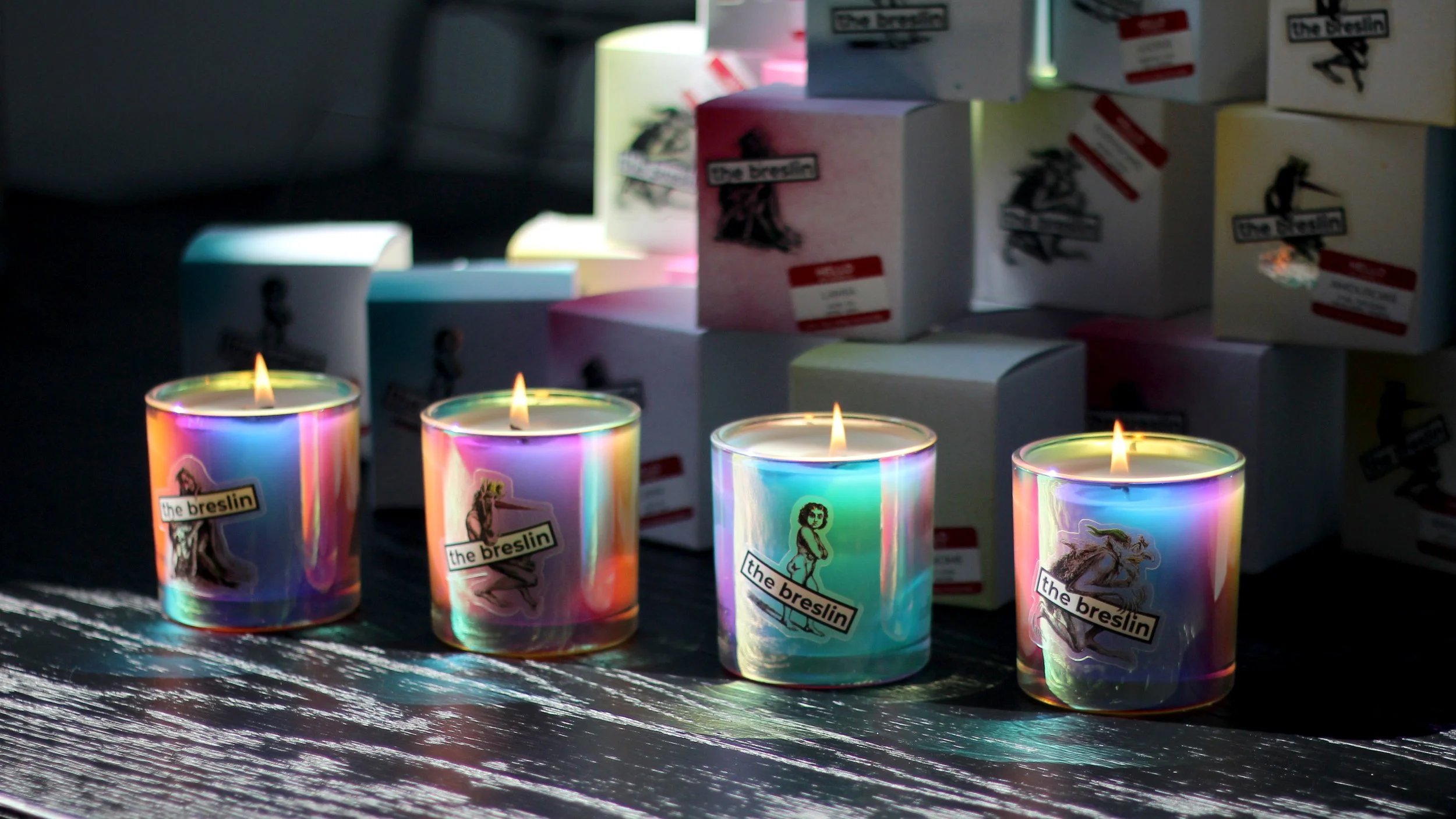 Four rainbow-colored candles with lit flames on a dark table, with boxes labeled 'The Breslin' in the background.