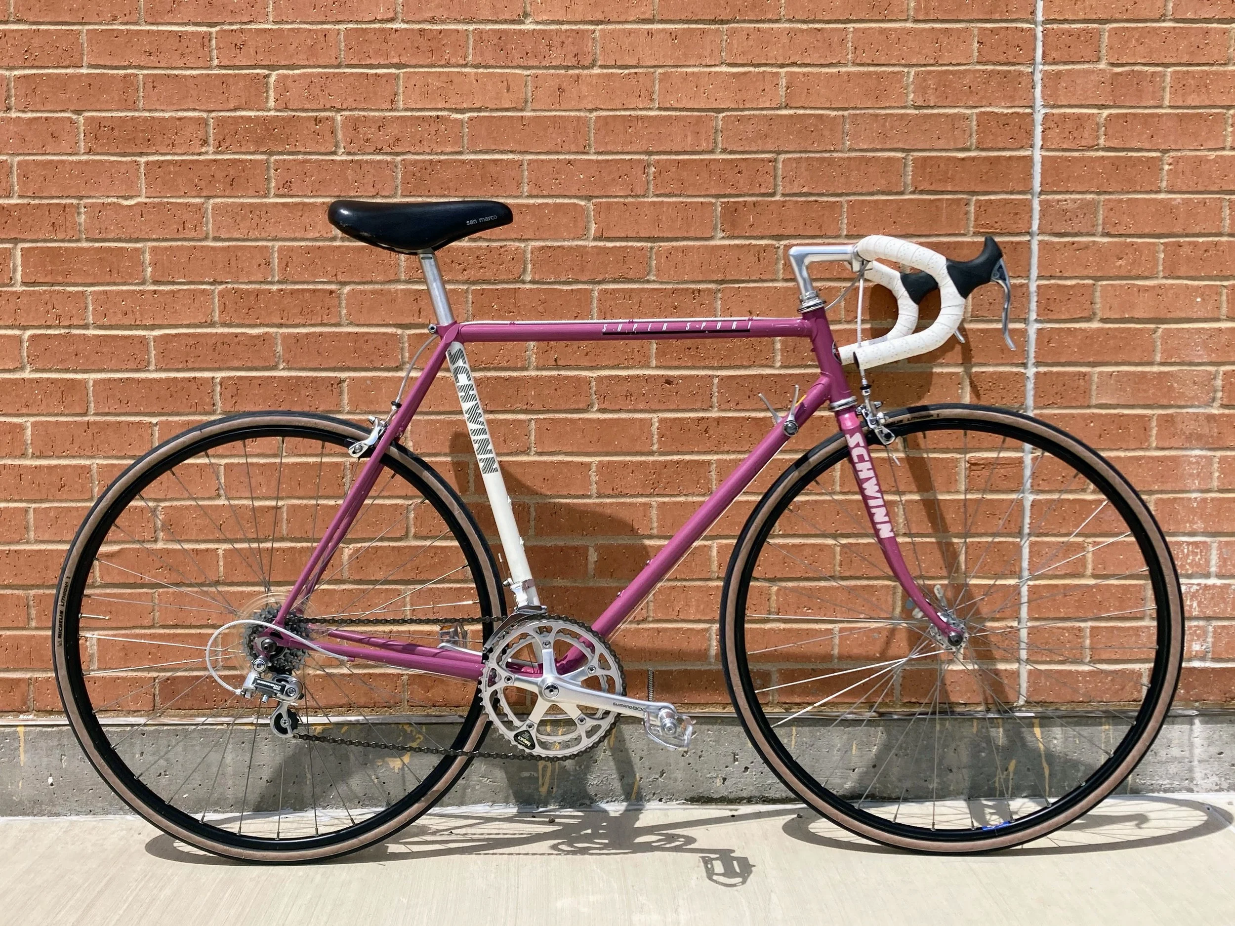A purple and white vintage Schwinn Super Sport road bicycle leaning against a brick wall.