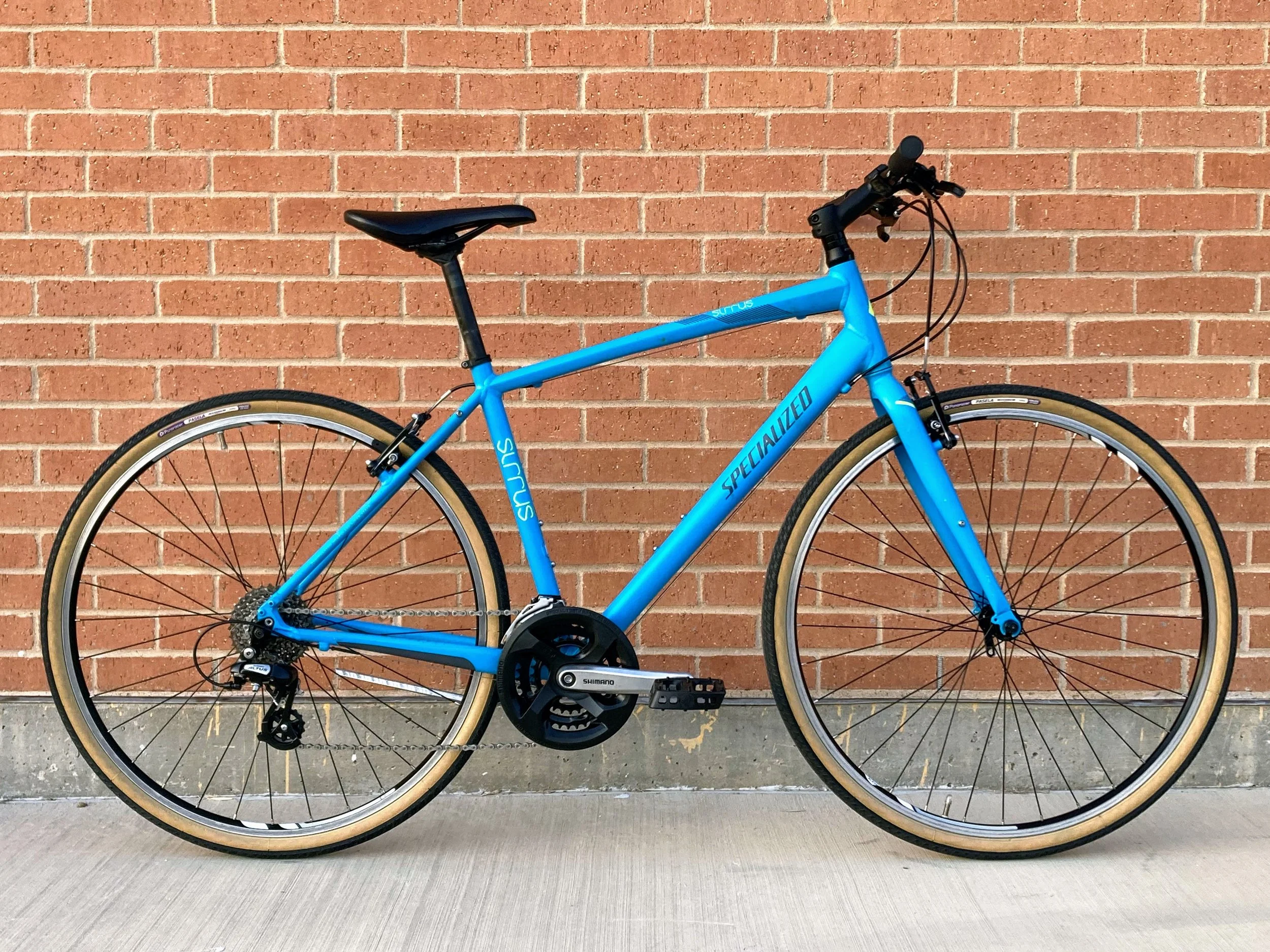A bright blue Specialized Sirrus hybrid bicycle with tan tires parked against a red brick wall.