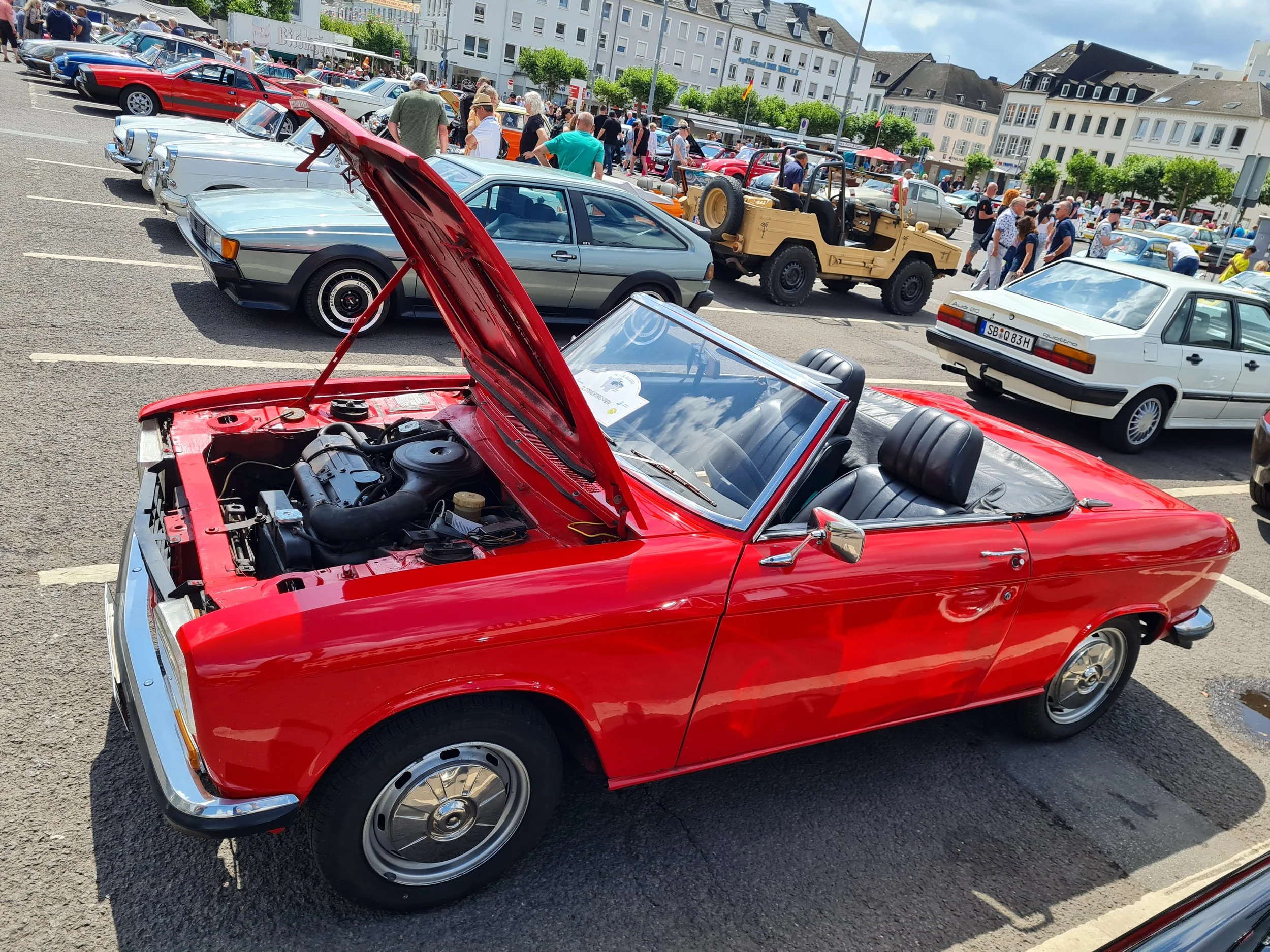 Rotes Cabriolet mit offenem Verdeck und geöffneter Motorhaube auf einem Parkplatz. Im Hintergrund sind weitere Oldtimer-Fahrzeuge und Menschen zu sehen, die an einer Autoausstellung teilnehmen.
