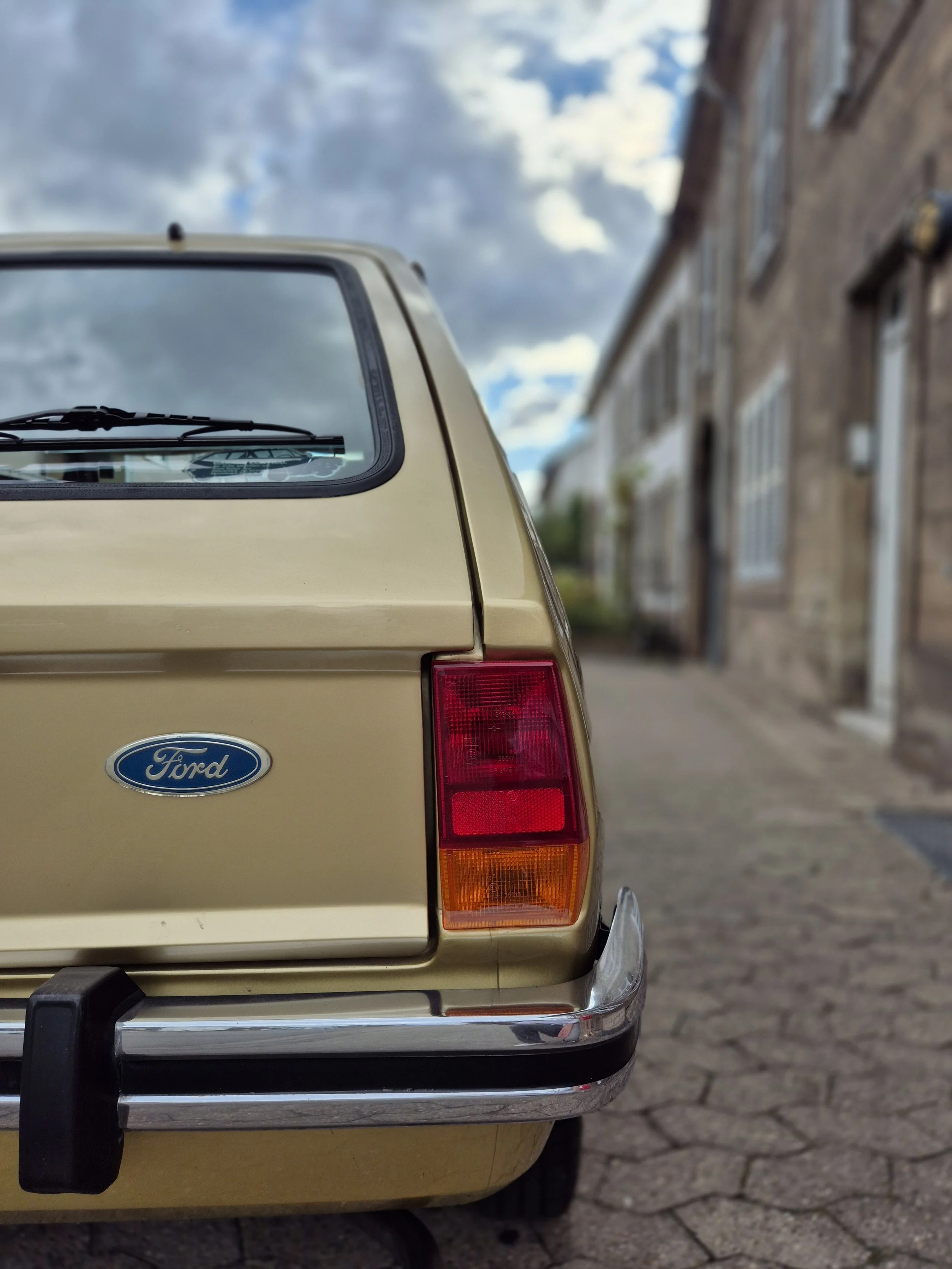 Rückansicht eines beigen Ford-Autos, geparkt auf einer Straße. Sichtbare Heckscheibe, Rücklicht und das Ford-Logo. Im Hintergrund unscharfe Gebäude und Wolken am Himmel.