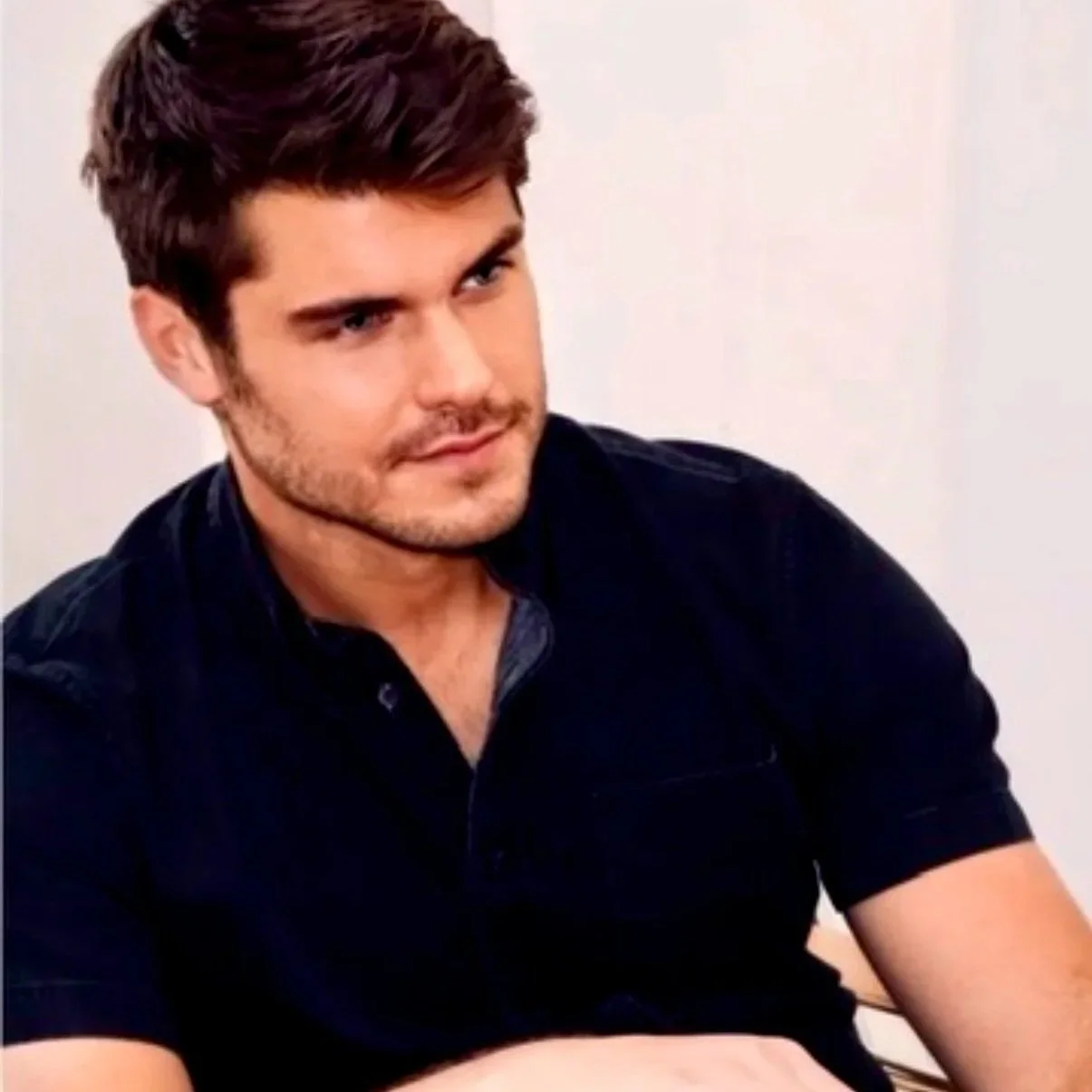 Young man with dark hair and beard wearing a black shirt, sitting against a plain white wall.