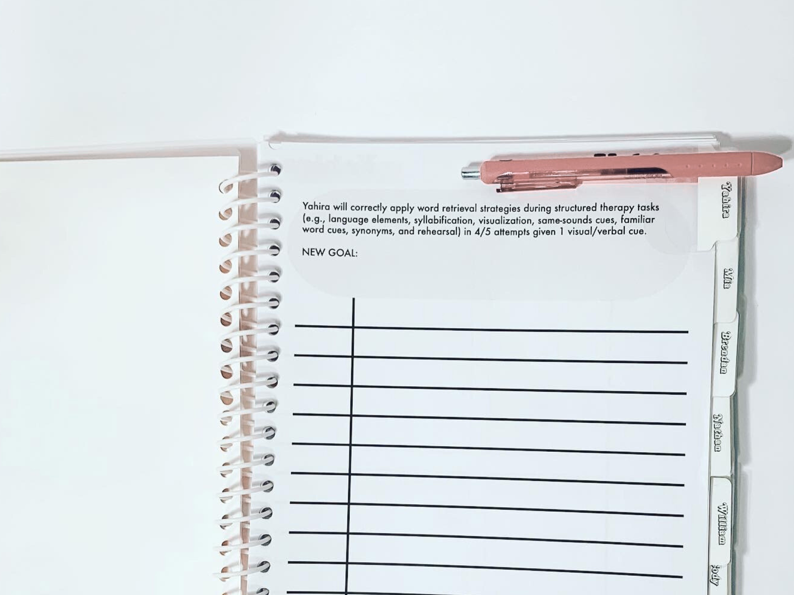 Spiral notebook with tabs labeled 'Yahya,' 'Eliya,' 'Brendan,' 'Nobia,' 'Daisy,' and 'Anah.' A pink pen is clipped to the top of the notebook, and a sheet of paper with printed text and blank lines is inside.