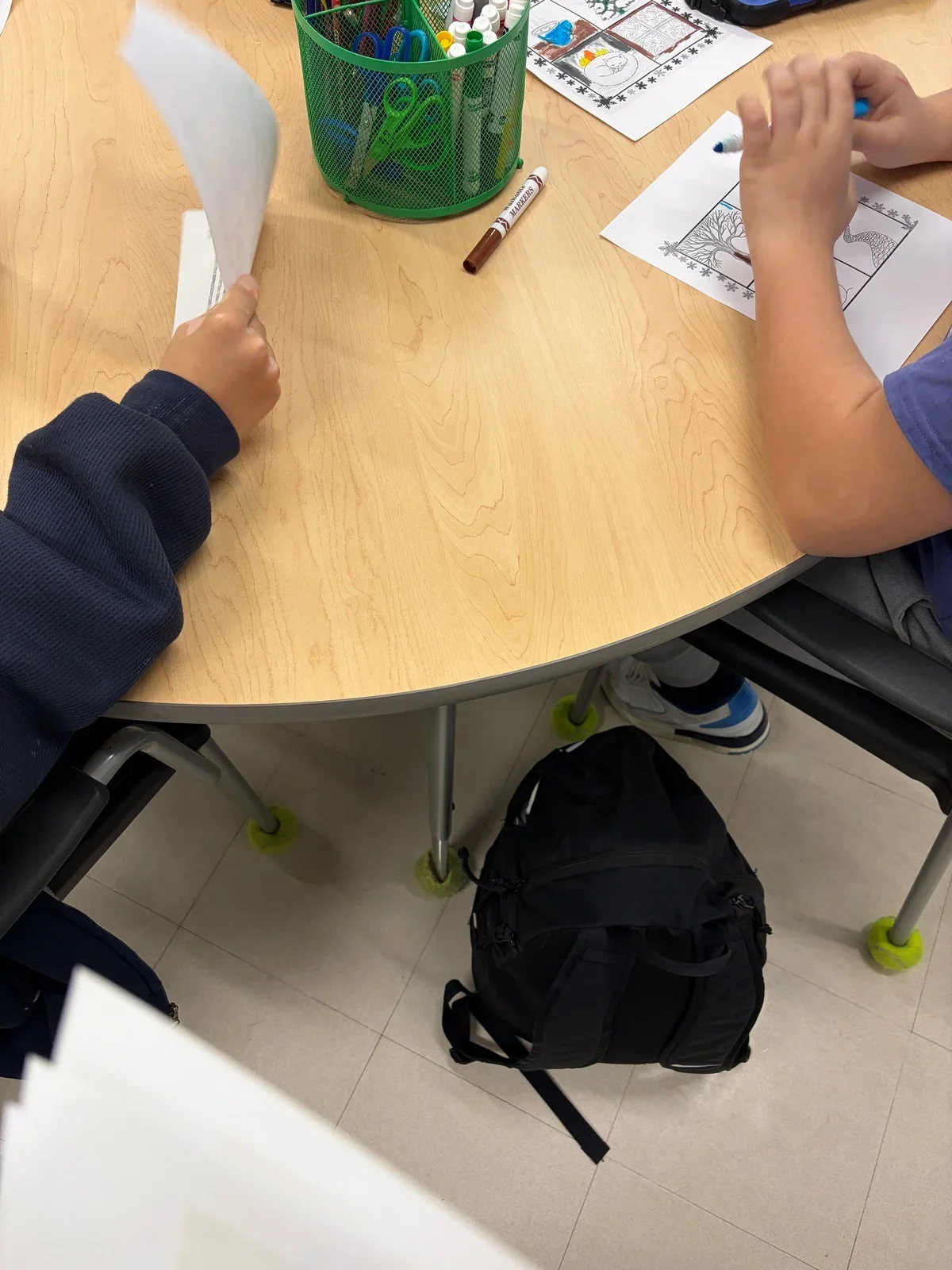 Students sitting at a classroom table, working on coloring sheets, with a green pencil holder filled with colored markers and scissors, a colorless marker, and a holiday-themed worksheet.