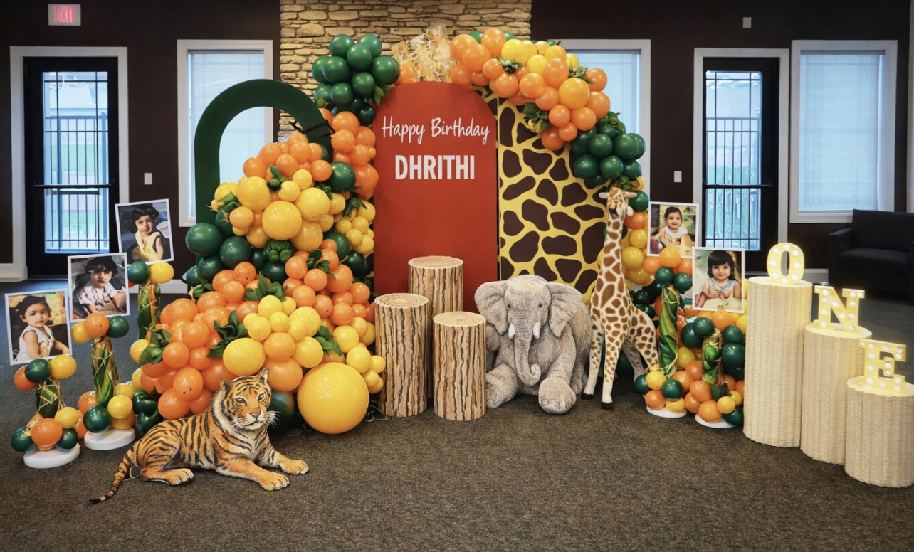 Safari-themed birthday party decoration with balloons, stuffed animal animals including a tiger, elephant, and giraffe, photo portraits of children, wood logs, and illuminated letters spelling "ONE".