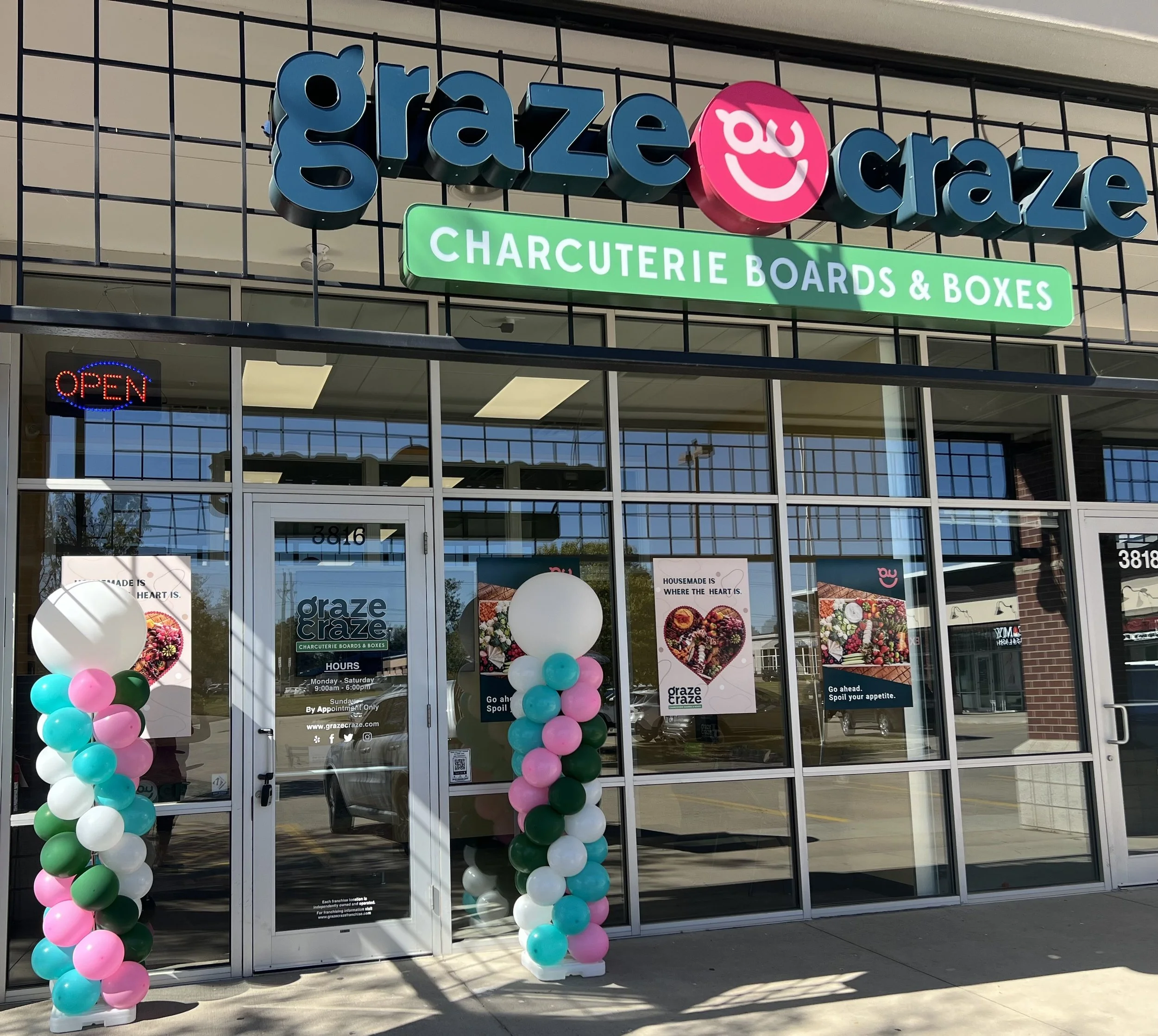 Front view of a storefront with a large sign saying 'graze craze' and a pink smiley face logo. Below, a green sign reads 'CHARCUTERIE BOARDS & BOXES.' An illuminated 'OPEN' sign is displayed in the window. Decorated balloon columns with pink, white, and green balloons are on either side of the entrance. Posters on the windows promote housemade food and encourage spoiling your appetite.