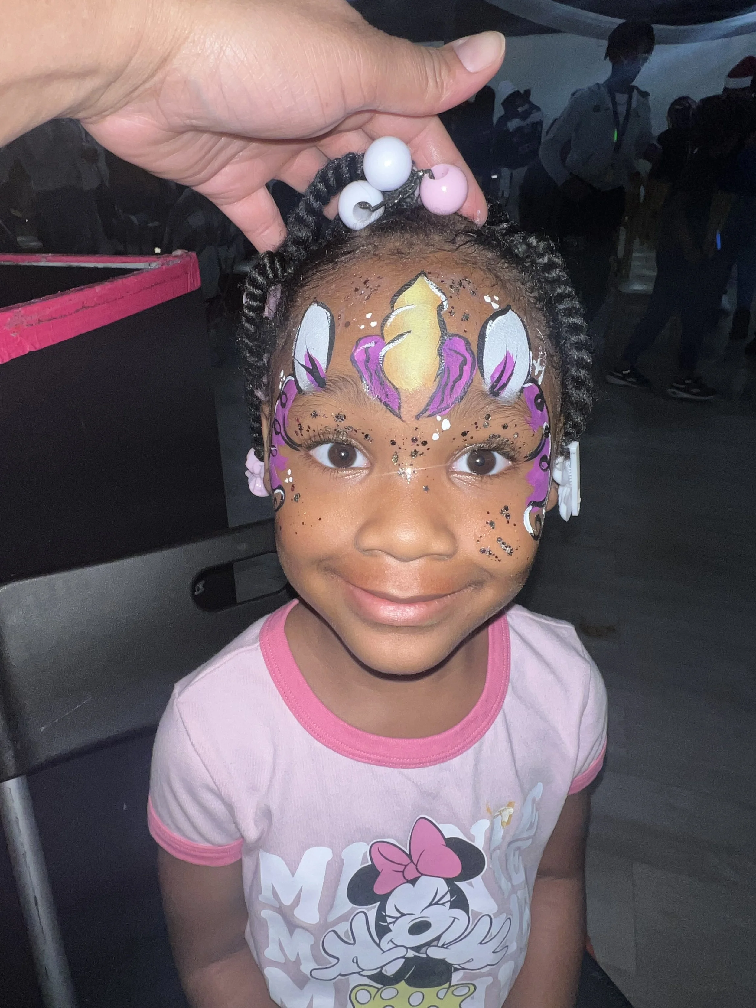 A young girl with face painting resembling a unicorn, highlighting her brown eyes and smiling face. A person's hand holds her head steady as the face painting is being finished. She is wearing a light pink t-shirt with Minnie Mouse on it.