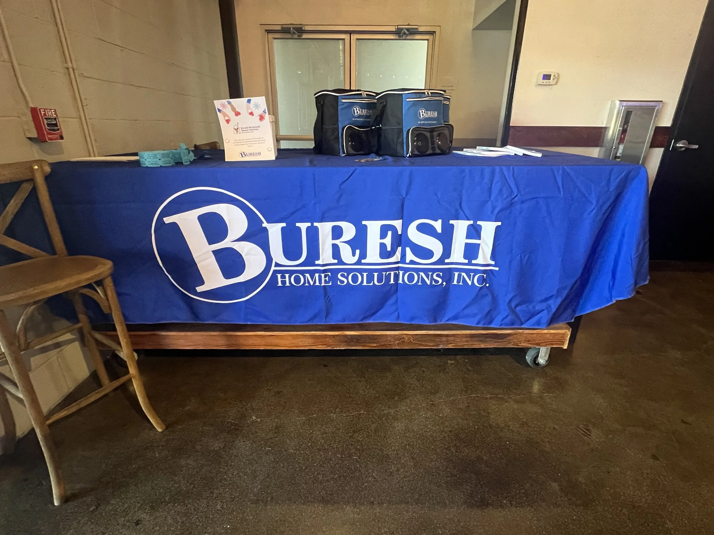 A table covered with a blue tablecloth displaying the Buresh Home Solutions, Inc. logo, with some promotional materials and bags on top, set up in an indoor space.
