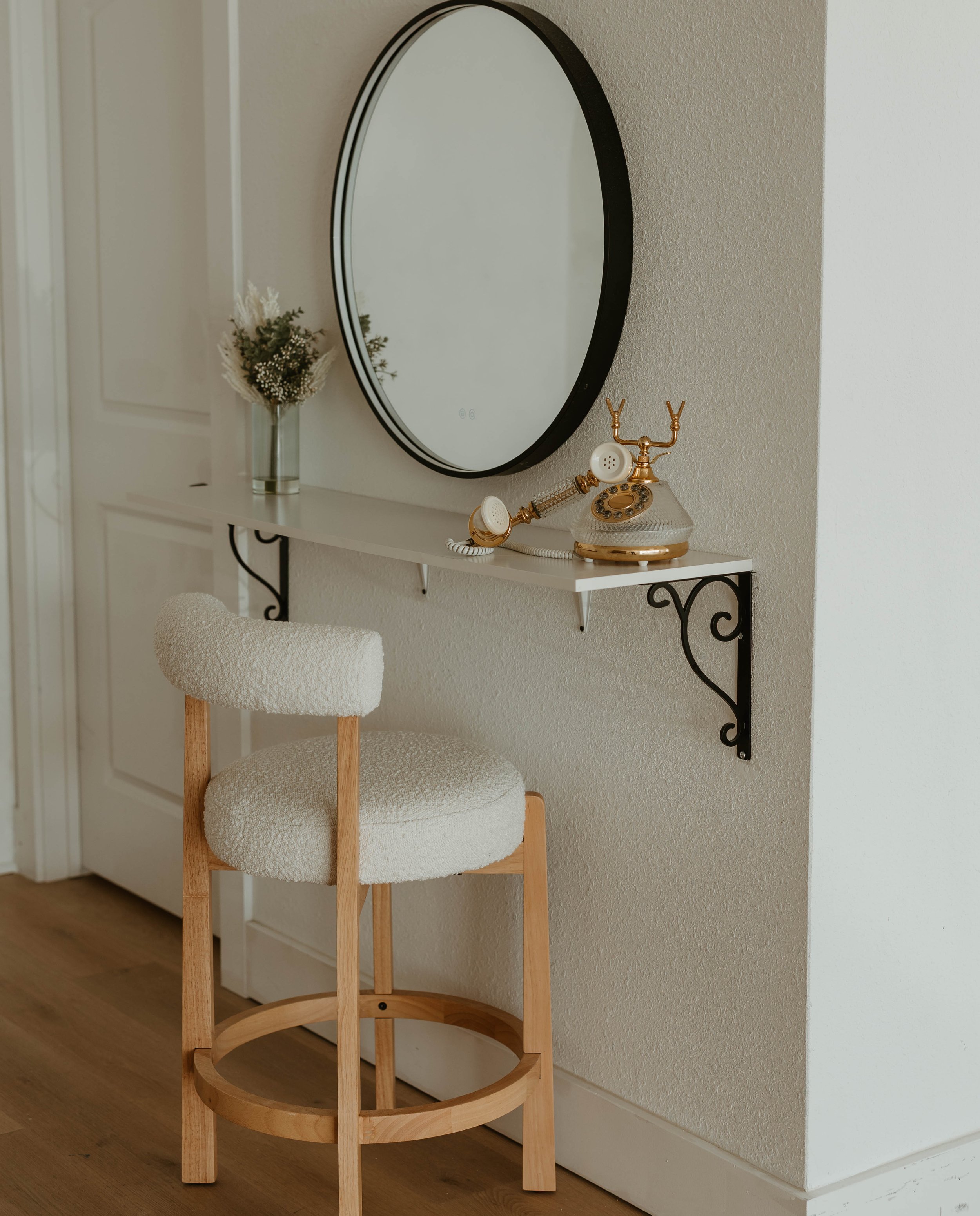 A room with a white wall, a hallway door, a window with light coming through, a round mirror on the wall, a small white shelf with black decorative brackets, a vintage telephone with a gold and glass design, a clear glass vase with dried flowers, and a wooden chair with a round, upholstered white seat and backrest.