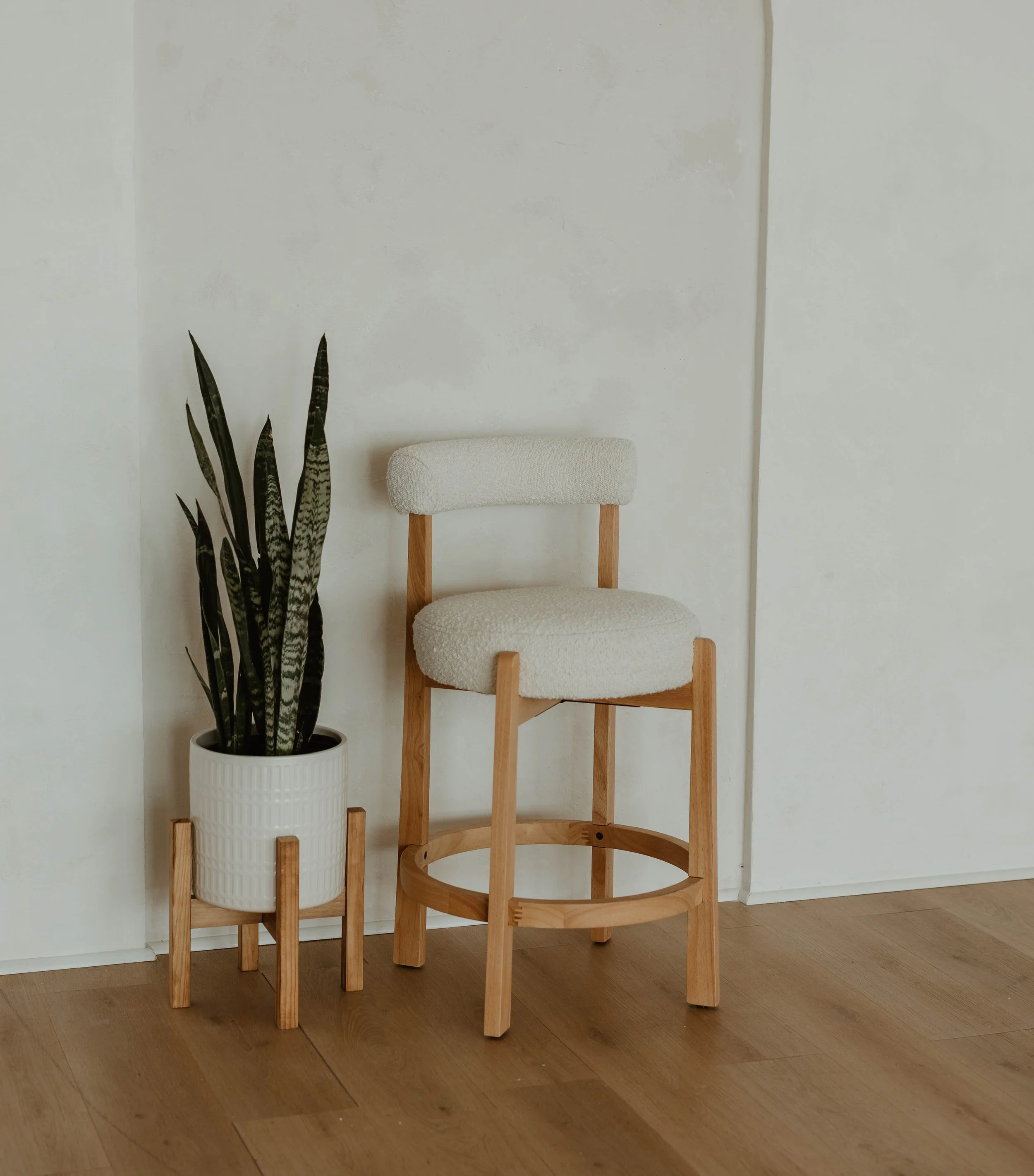 A white upholstered chair with wooden legs and a circular frame base, next to a potted snake plant in a white textured pot with a wooden stand, against a white wall.