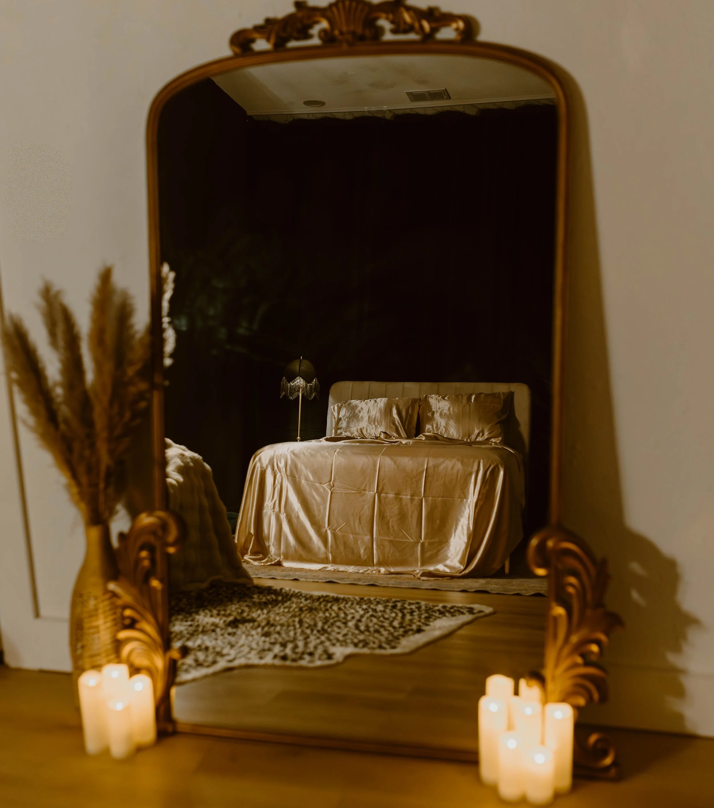 A mirror reflecting a bedroom with a bed covered in a shiny beige or gold silk comforter, matching pillows, a black nightstand with a lamp, a black wall, and a patterned rug. Candles are placed at the base of the mirror.