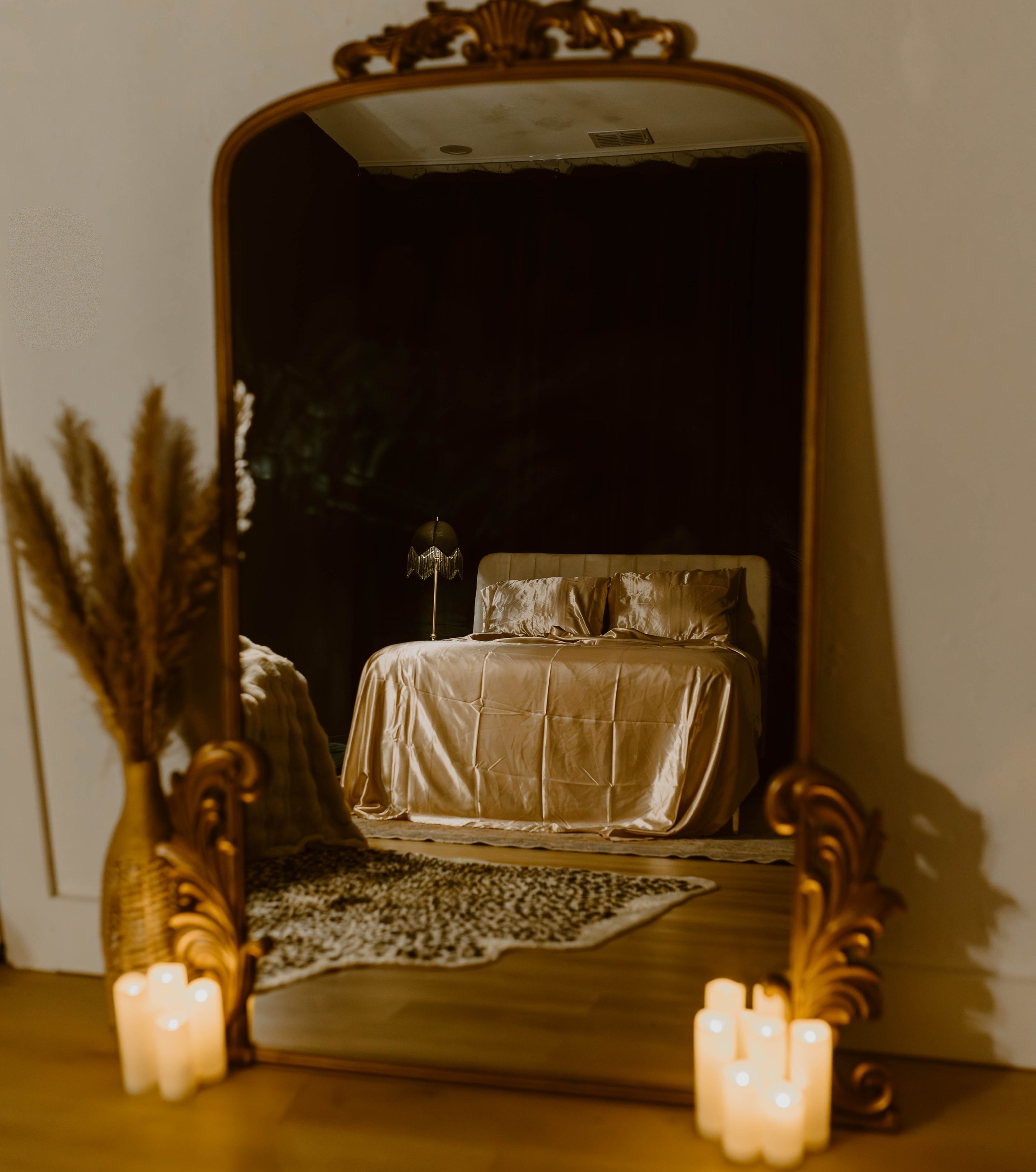 A bedroom seen through a large ornate mirror leaning against a wall, with candles lit on the floor around it. The bed has satin bedding and a black lamp beside it.