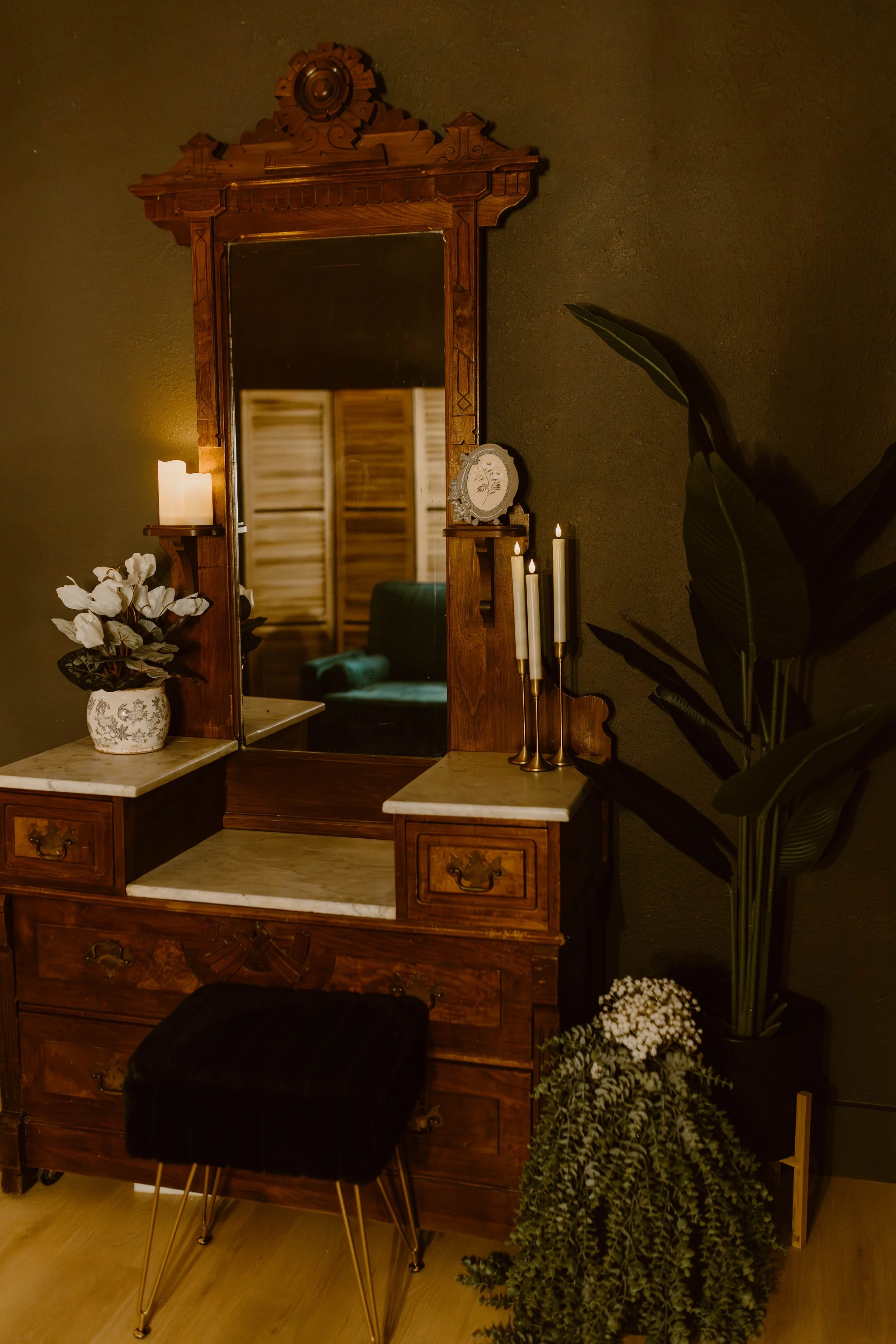 Vintage wooden dresser with a mirror, decorated with white flowers in a patterned vase, candles in gold holders, and framed pictures, set against a dark wall with large green indoor plants.