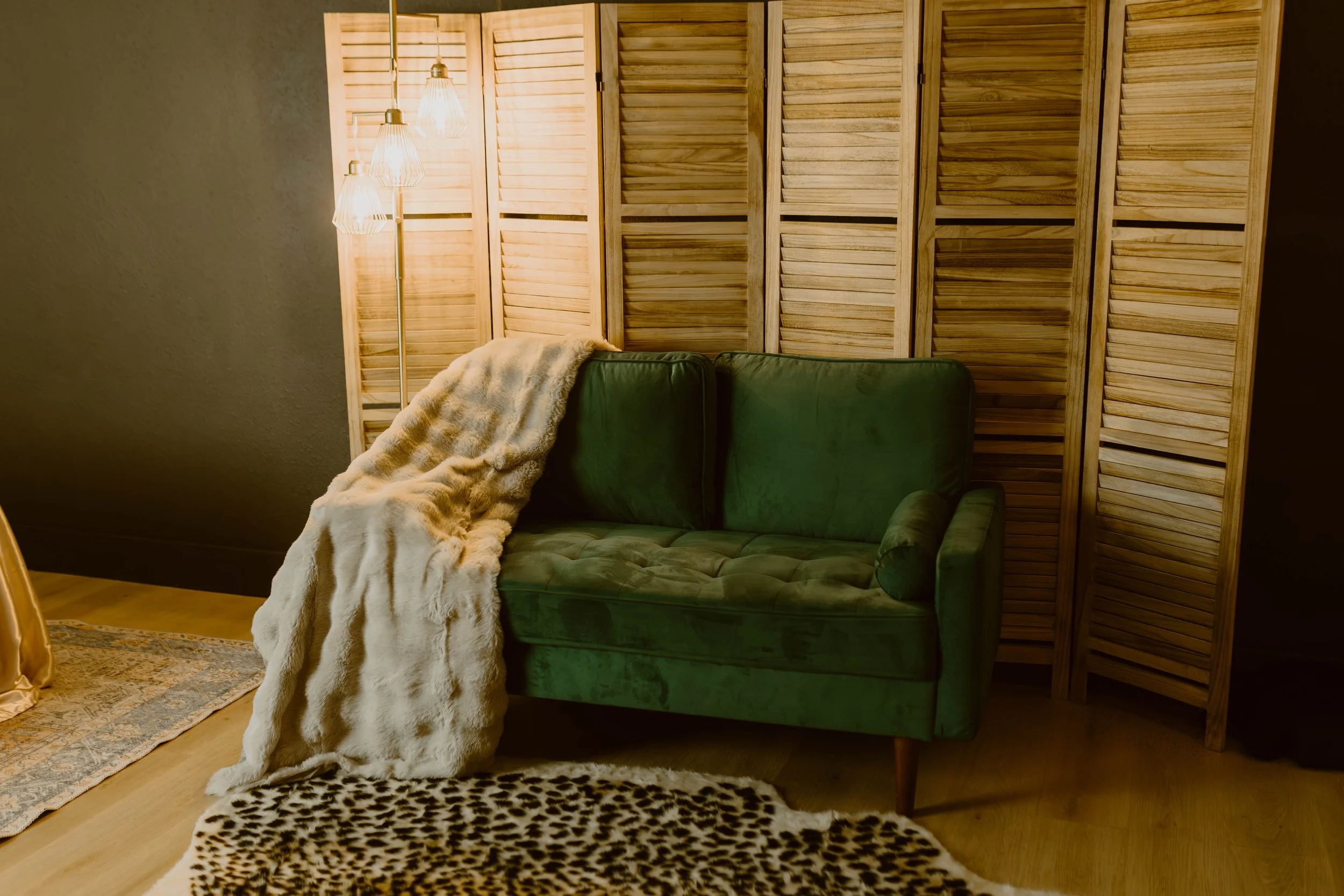 A vintage green velvet sofa with a rolled armrest, placed in a room with wooden flooring and a wooden privacy screen. A faux fur throw blanket is draped over the sofa's left arm. To the left, there is a standing floor lamp with three warm-colored han