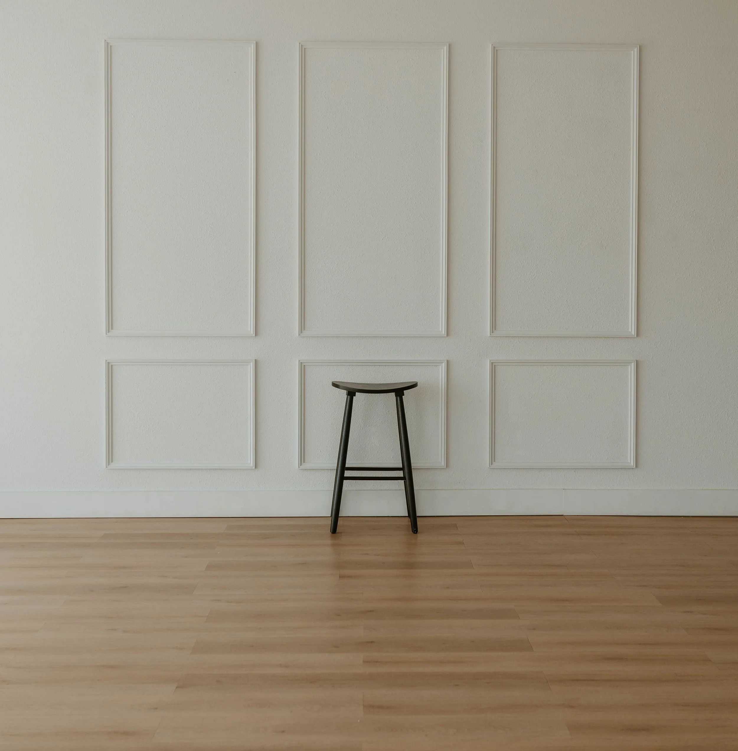 A minimalistic room with white paneled walls, light-colored wooden flooring, and a black stool in the center.