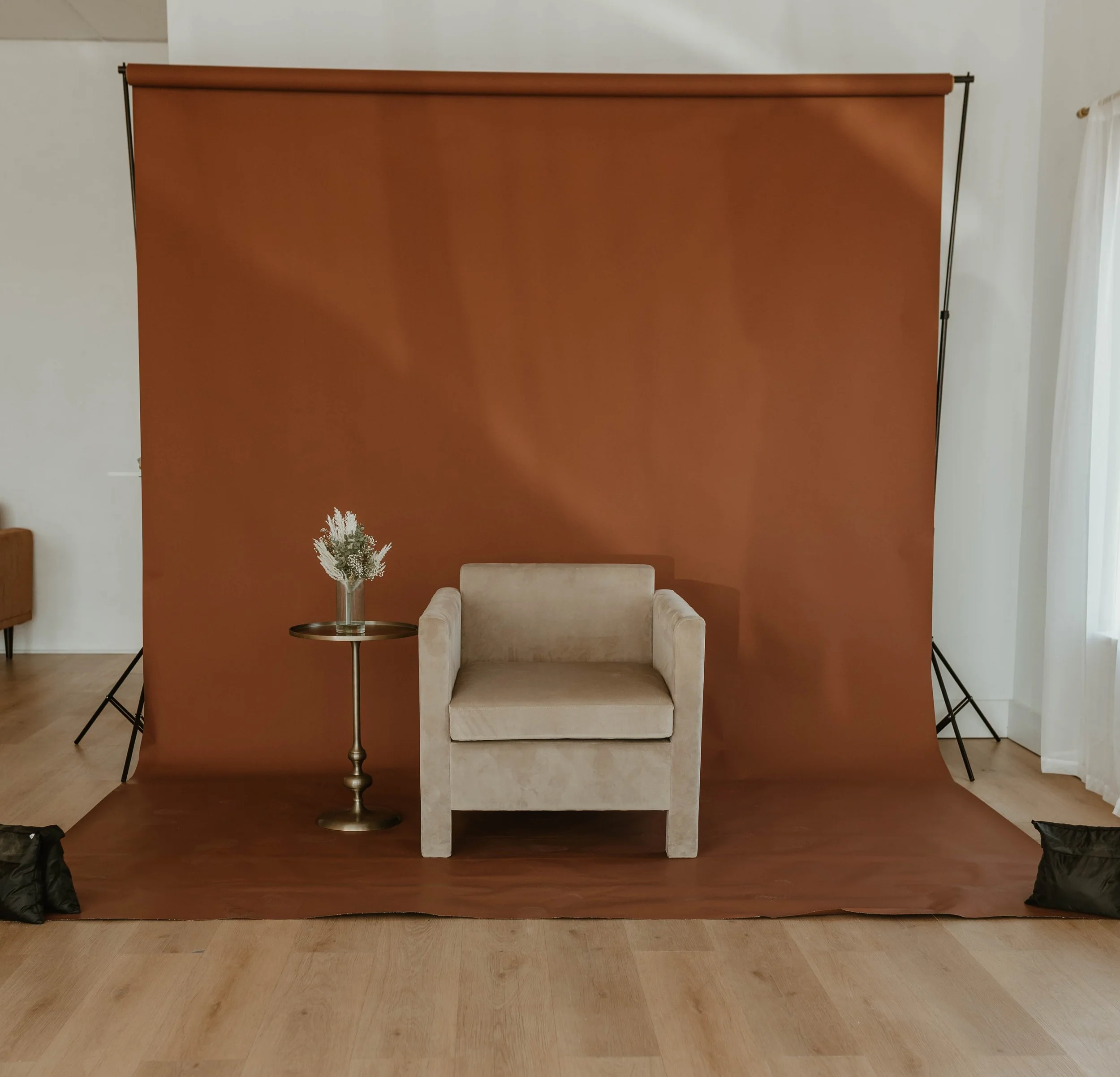 A beige armchair in front of a brown backdrop with a small round side table holding a flower arrangement to the left of the chair.