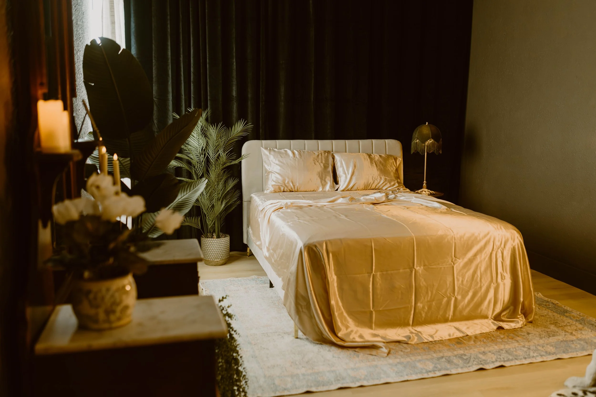 A bedroom with a cream-colored bed dressed in shiny golden bedding and pillows. A black wall behind the bed with dark curtains, and a bedside table with a vintage brass lamp. Plants in pots are near the window.