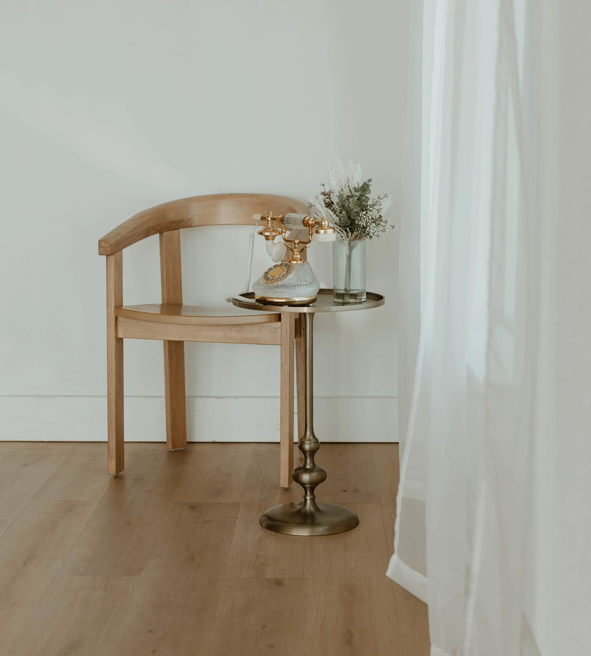 A vintage rotary telephone and a glass vase with eucalyptus and white flowers on a small round metallic side table next to a wooden chair with a curved back, in a room with light-colored walls, wooden floor, and sheer white curtains.