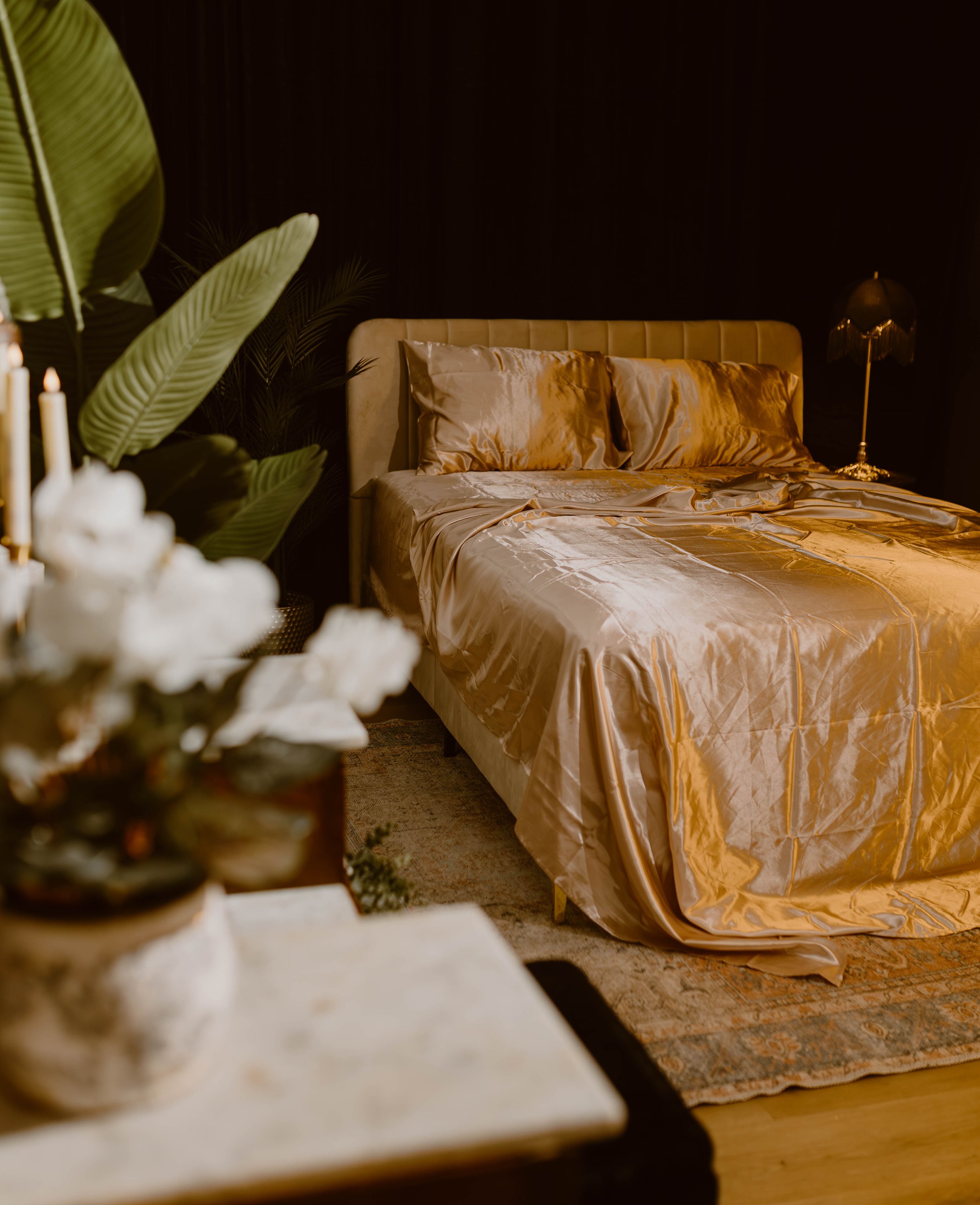 Luxurious bedroom with a gold satin bedspread and pillows, dark wall, gold-colored bedside lamp, and greenery in the background.