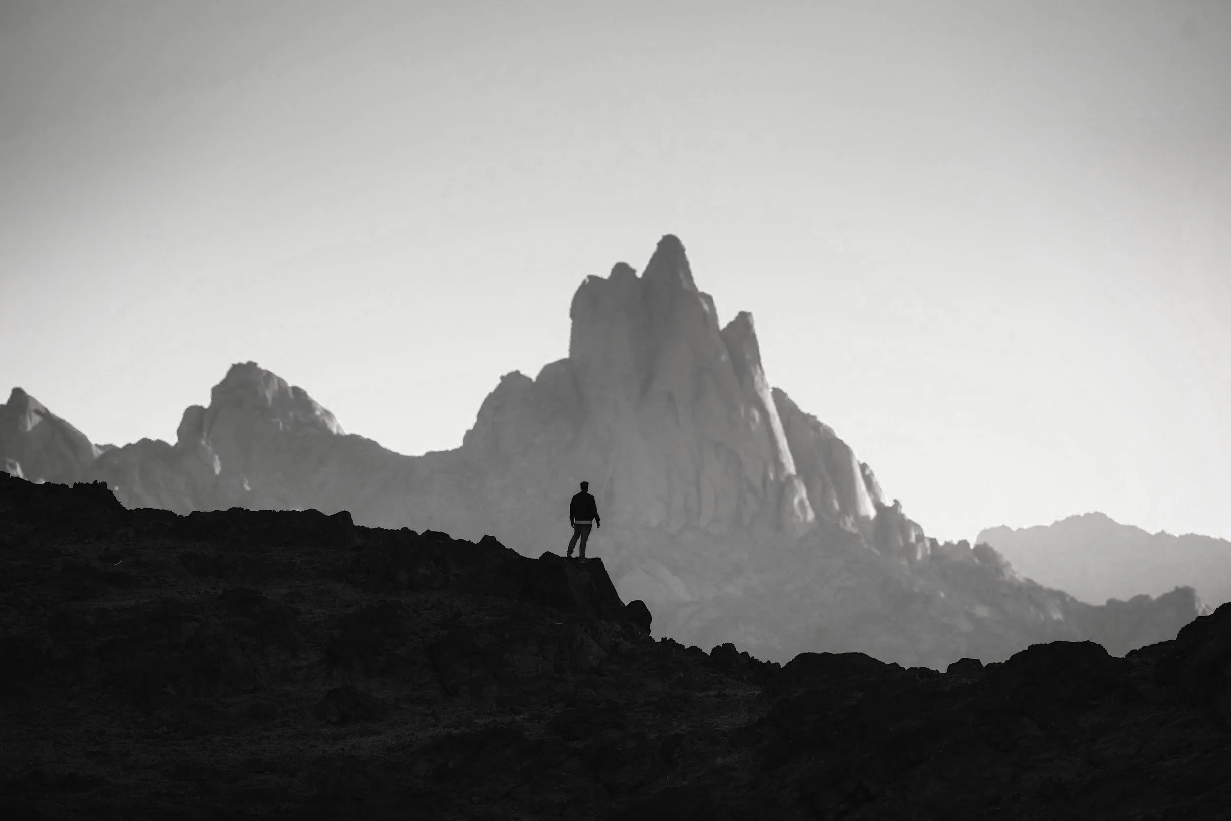 A person standing on a rocky hill silhouetted against a large mountain range in the background.