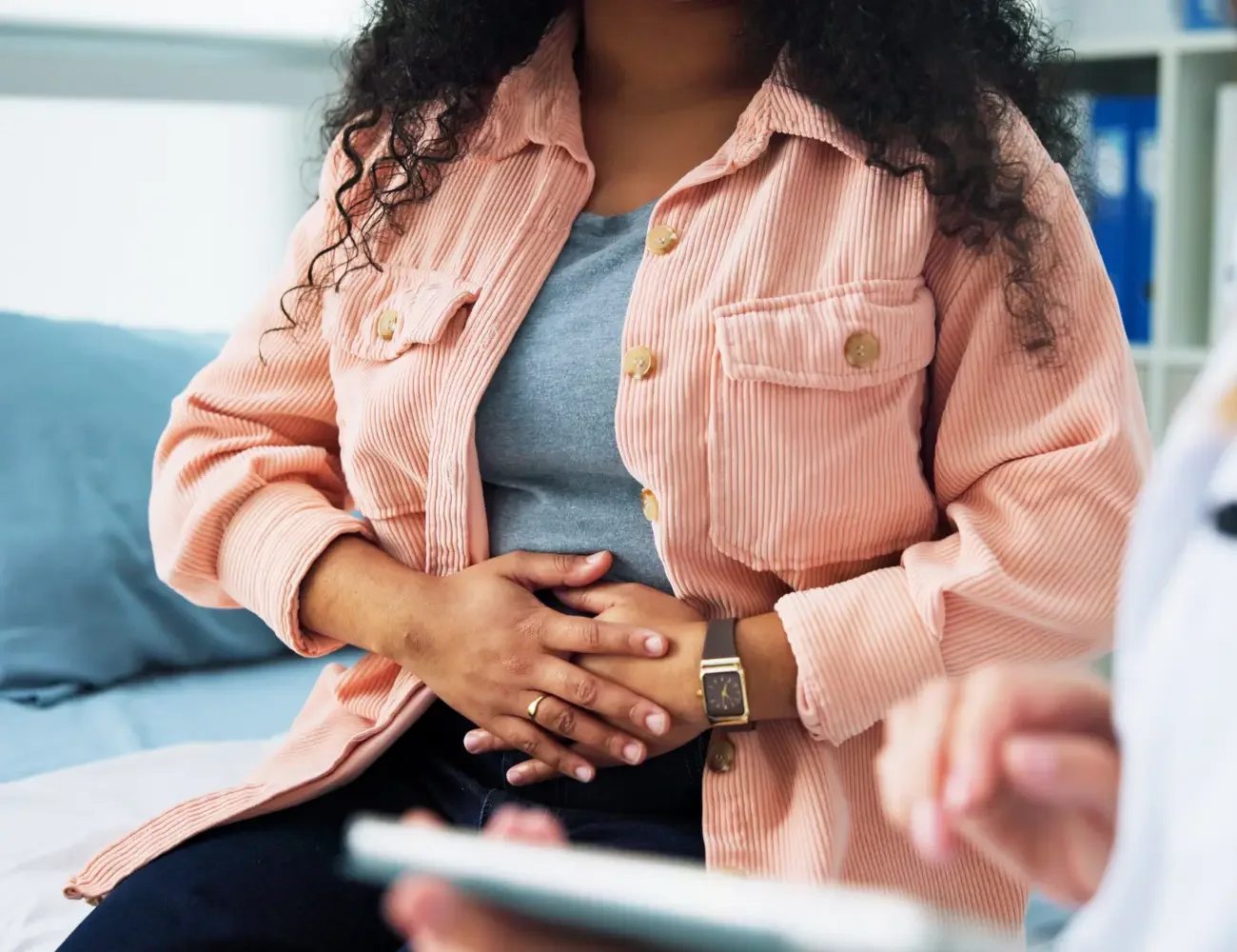 A woman sitting in a chair, holding her stomach with a pained expression, indicating discomfort or distress.