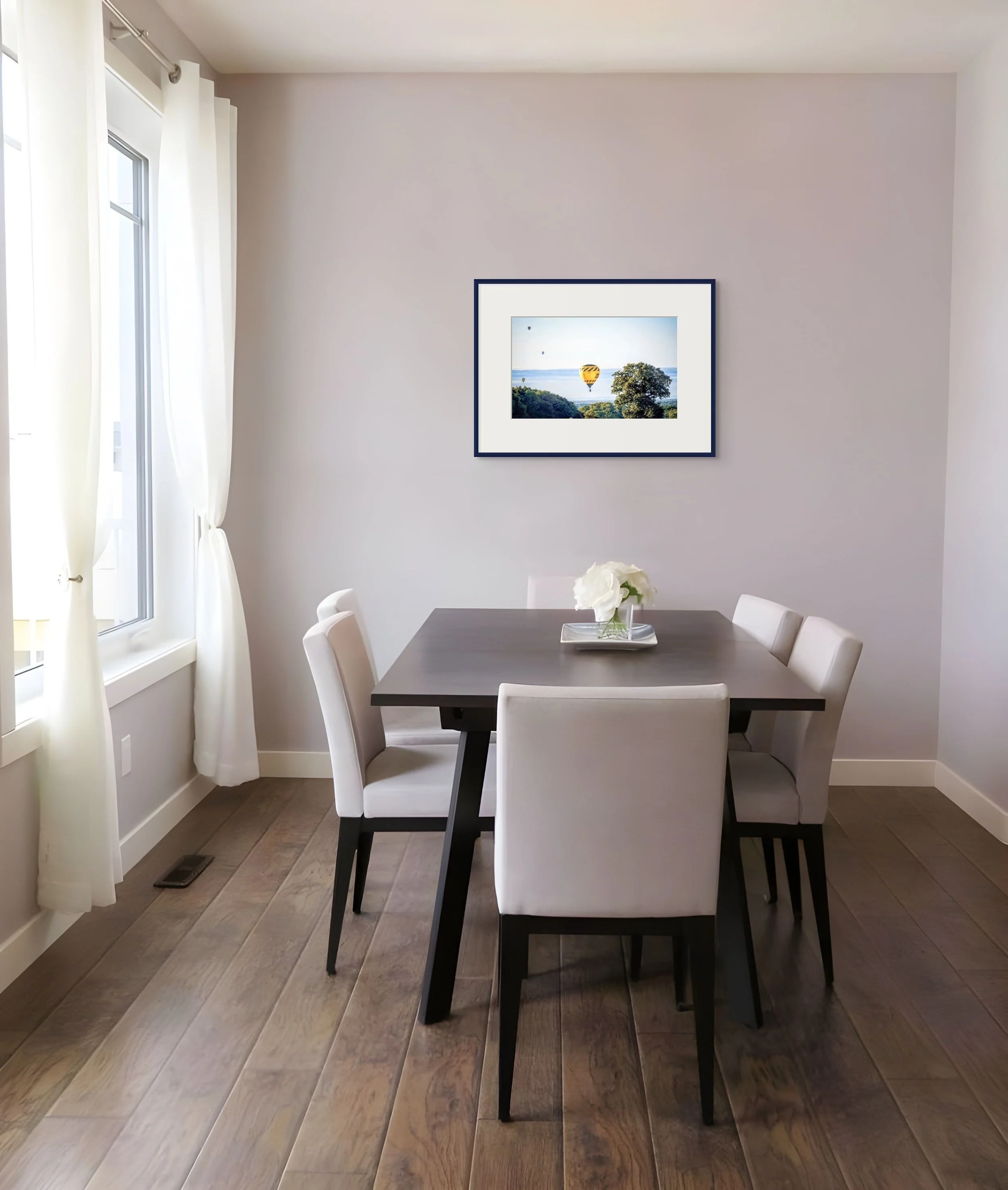Framed photo of a yellow hot air balloon floating above green hills and trees, hung above a dining table with white flowers. A quiet rural scene that hints at gentle movement and morning light.