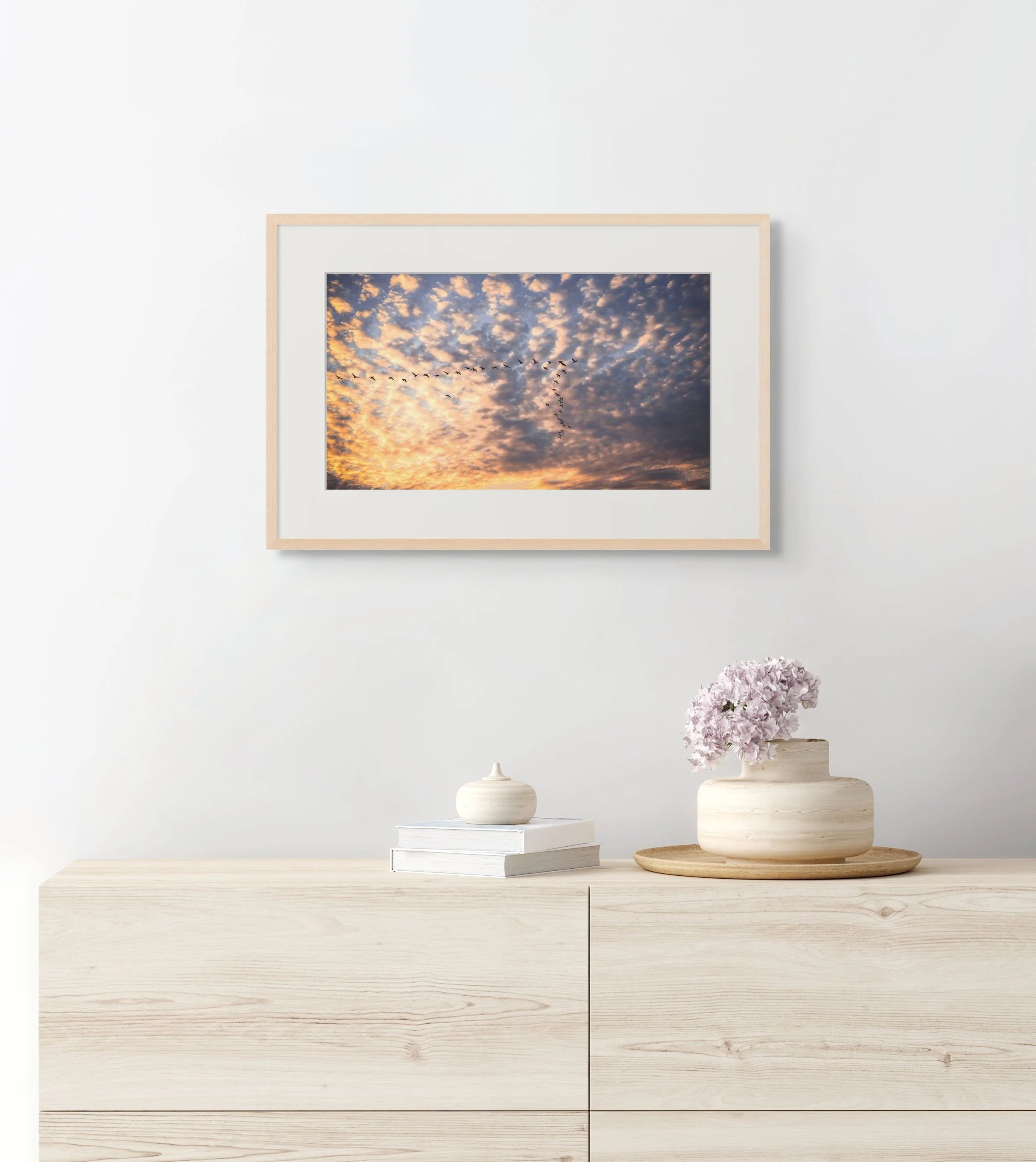Framed photo of birds in V-formation at sunrise hangs above a dresser with books, flowers, and ceramic decor in a serene interior setting.