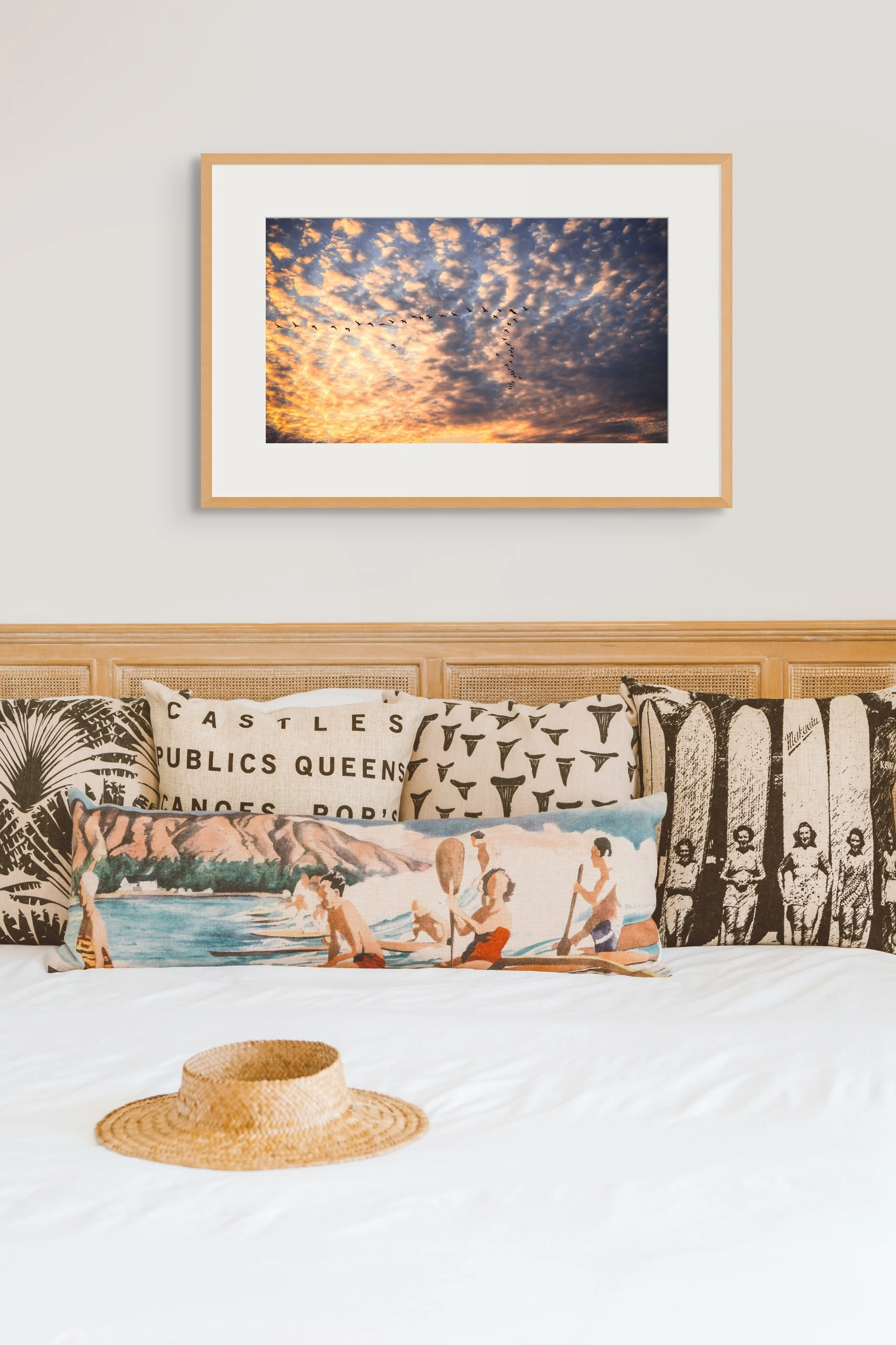 Cozy bedroom with eclectic pillows, a straw hat on white bedding, and a framed sunrise photo of birds in flight above the bed.