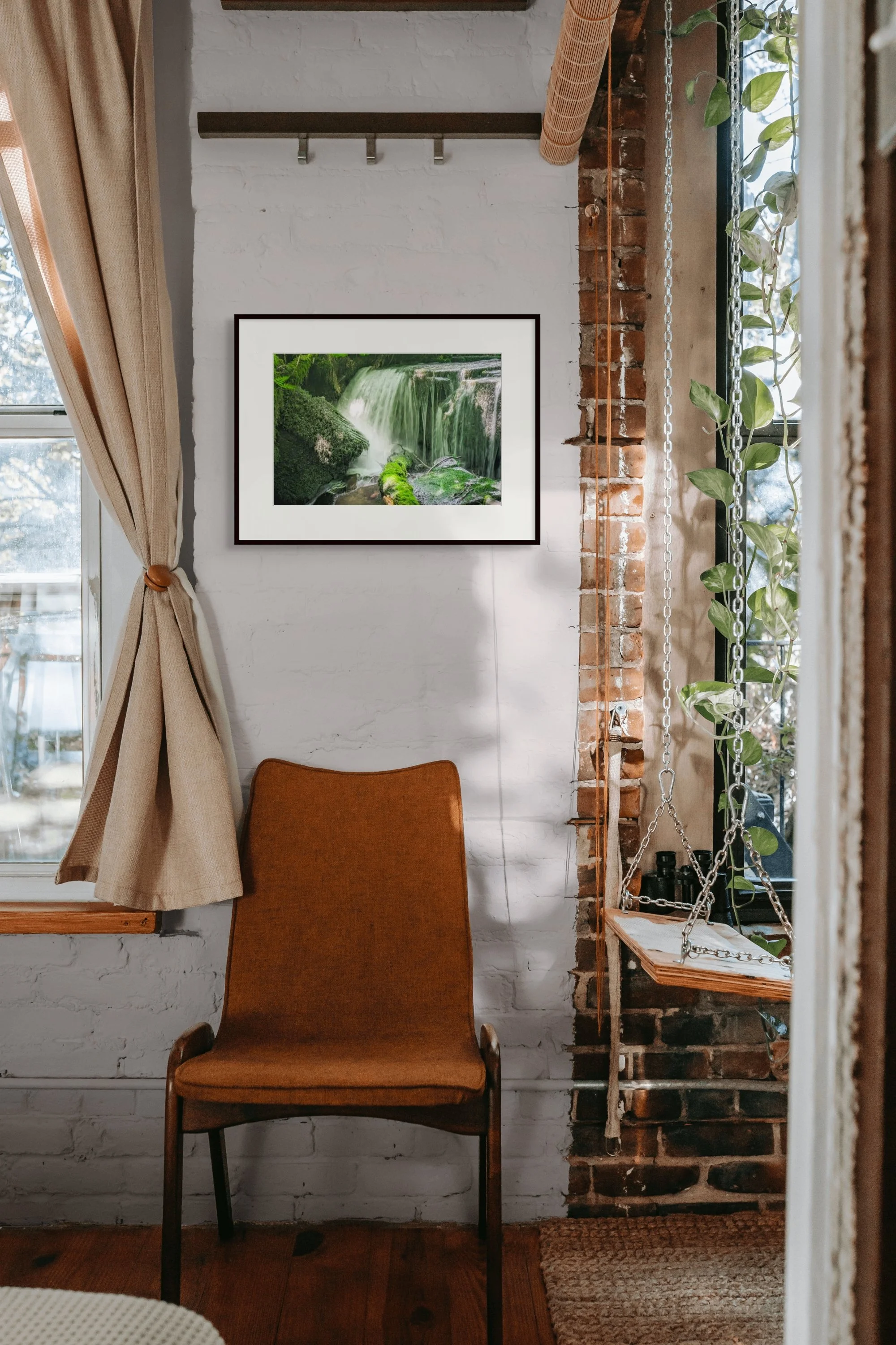 Cozy room corner with brown chair, framed waterfall photo, white brick wall, natural light from window, and rustic accents like wood and greenery.