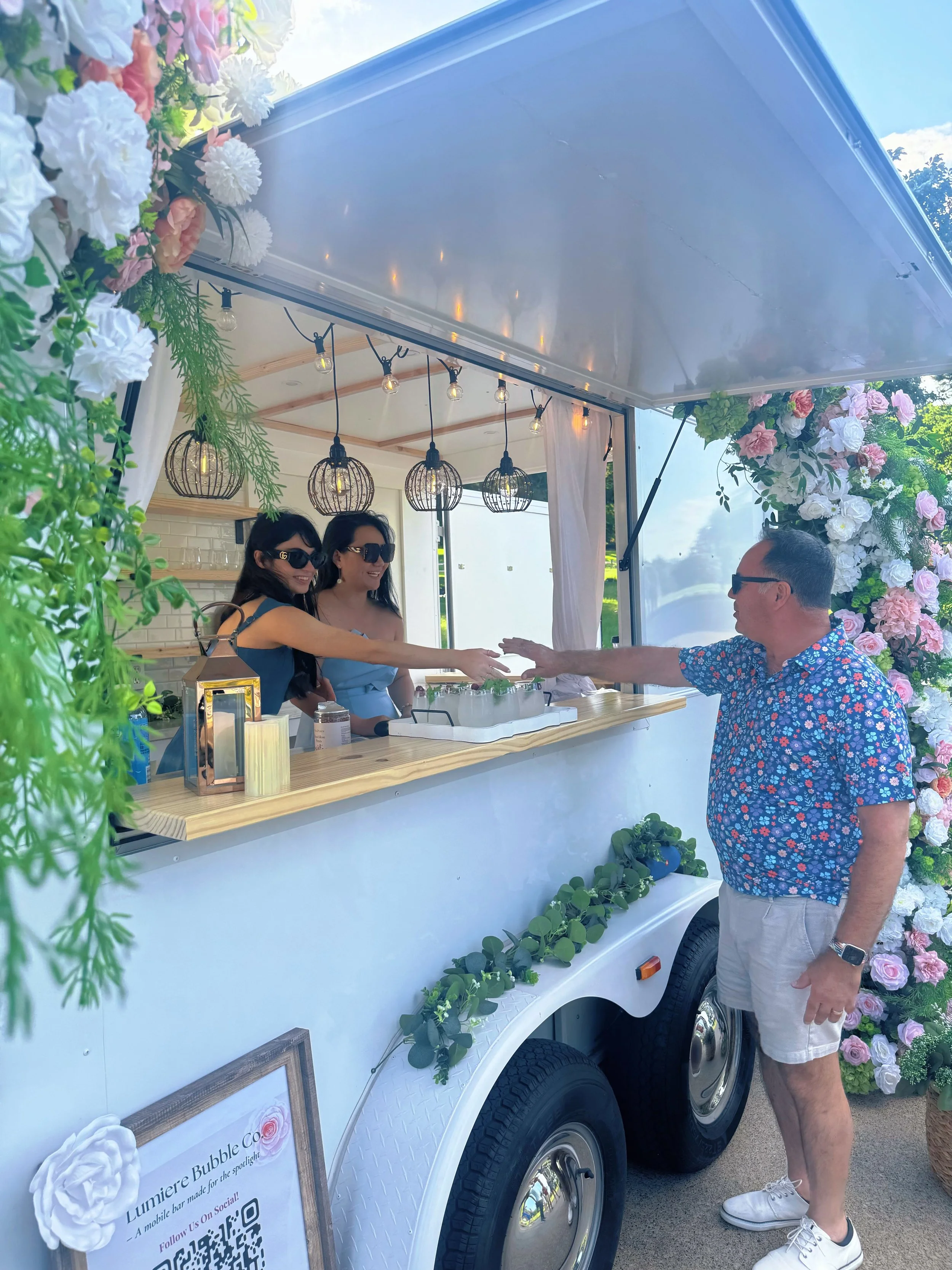Lumière Bubble Co. bartenders serving a guest from a boutique mobile bar trailer at an outdoor Dallas-Fort Worth event.