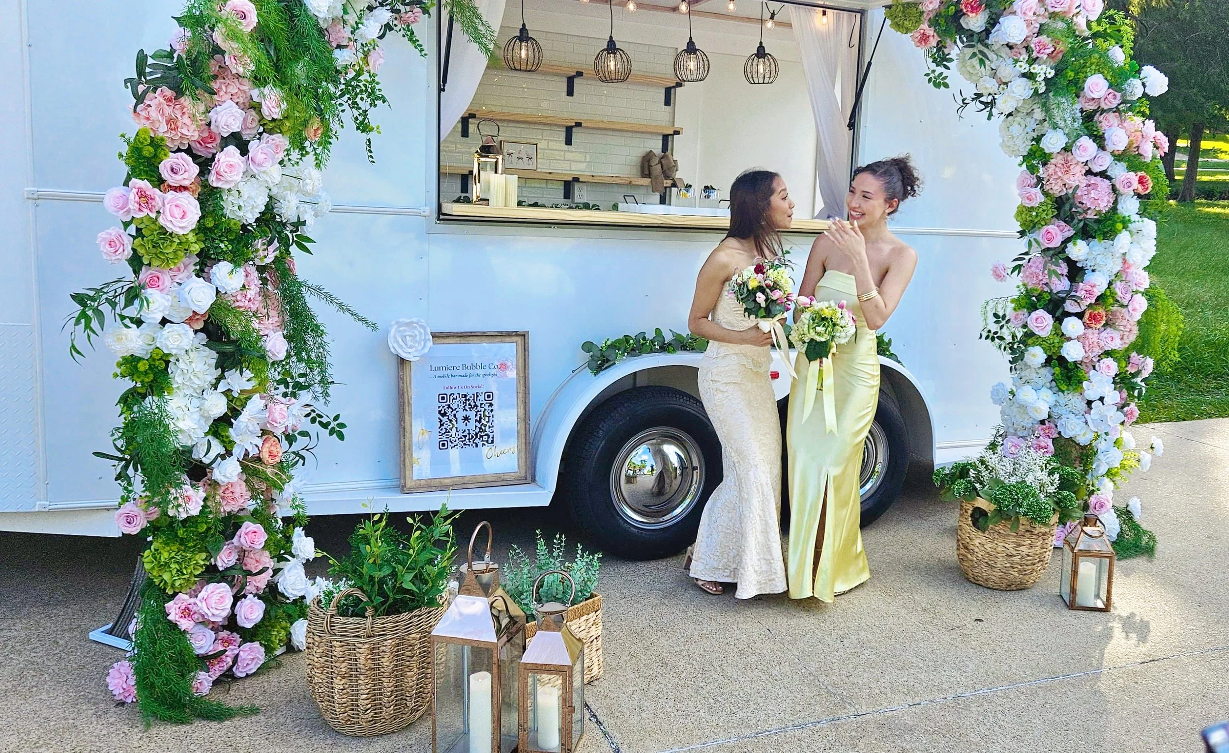 Two guests in their beautiful prom dresses holding floral bouquets posing in front of Lumière Bubble Co.'s pink-and-white floral mobile bar trailer at an outdoor Dallas-Fort Worth celebration.