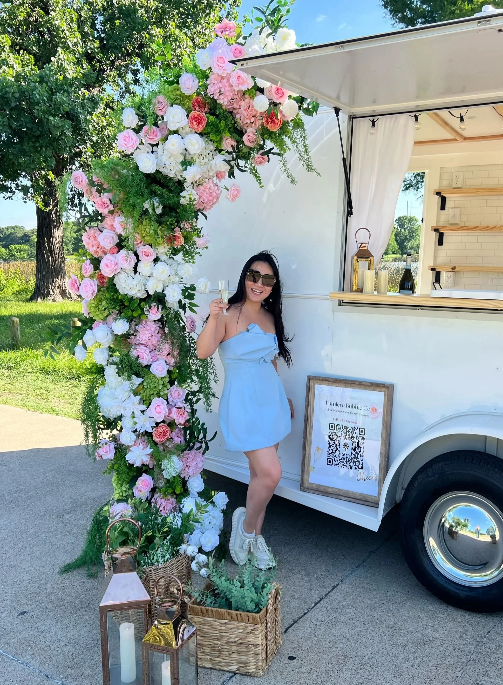 A woman in a blue dress, wearing sunglasses and sneakers, holding a champagne glass and smiling, standing next to a flower arrangement attached to a food truck. The truck has a sign with a QR code and the name 'Lumiere Bubble Co.' in front of a green outdoor setting with trees and a field.