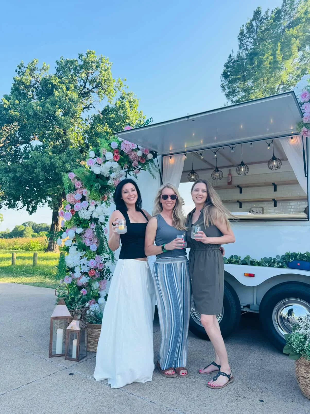 Three guests holding signature cocktails beside Lumière Bubble Co.'s floral mobile bar trailer at an outdoor Dallas-Fort Worth event.