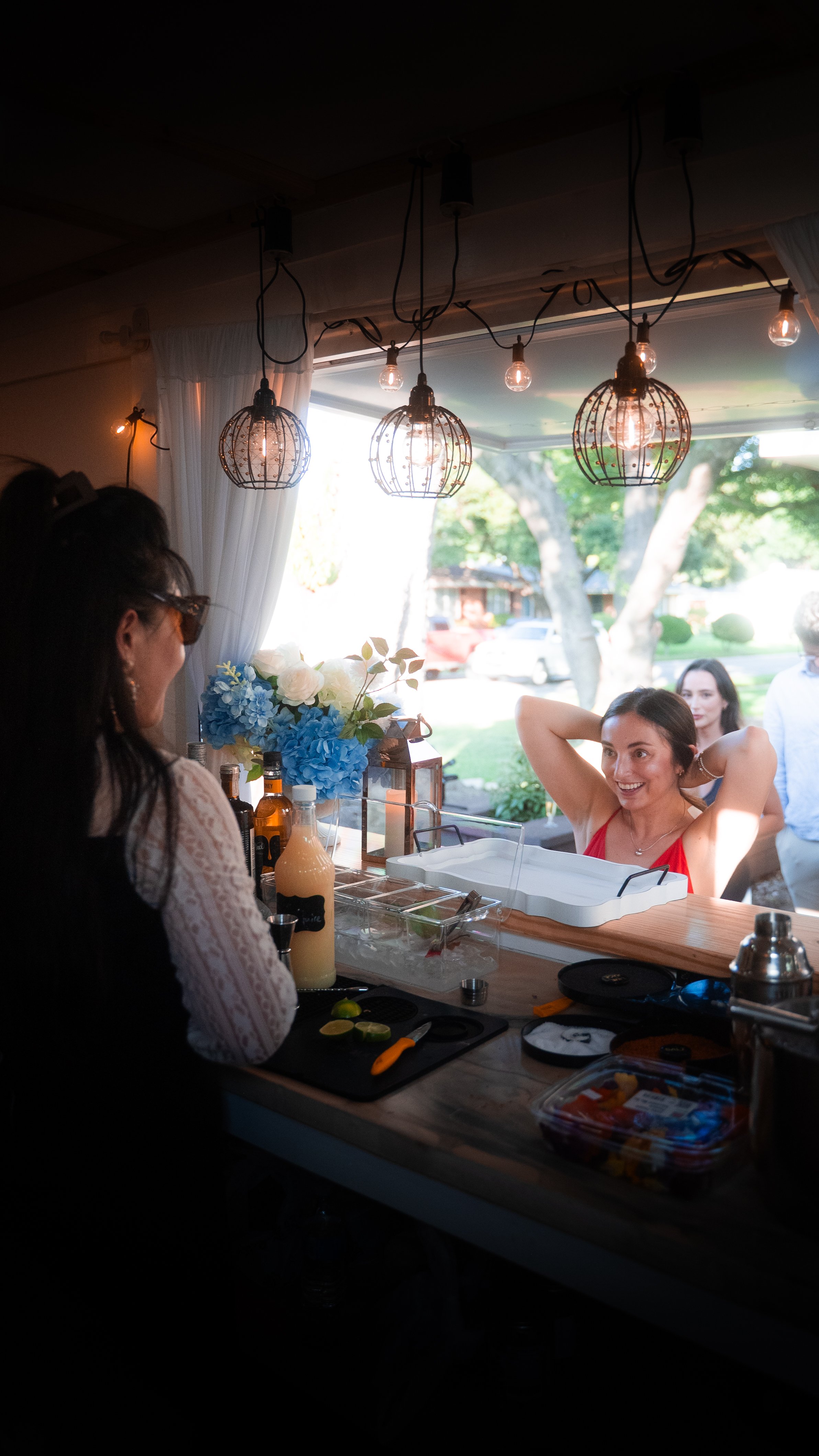 People preparing food at a bar with a woman in a red dress smiling with her hands behind her head, outdoor scenery visible through large windows.