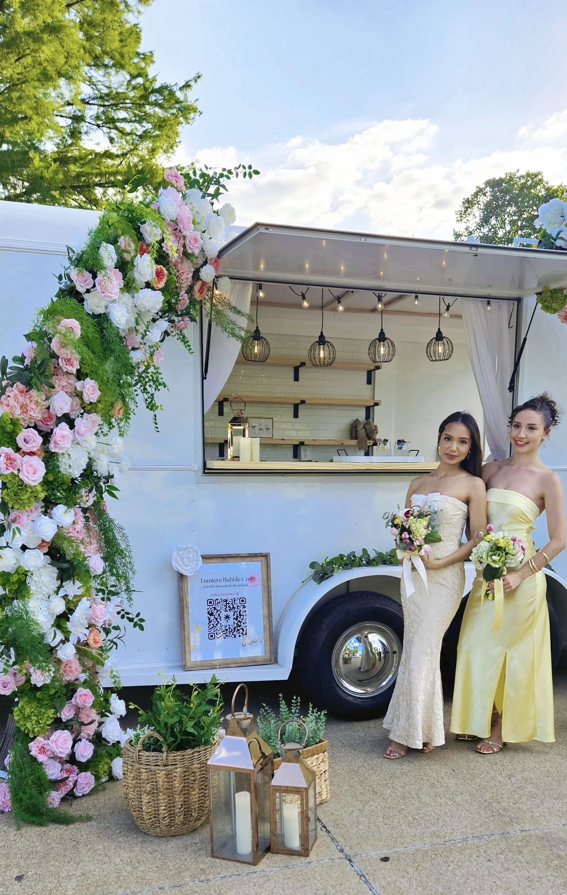 Two guests holding floral bouquets beside Lumière Bubble Co.'s flower-covered luxury mobile bar trailer at a Dallas-Fort Worth bridal event.