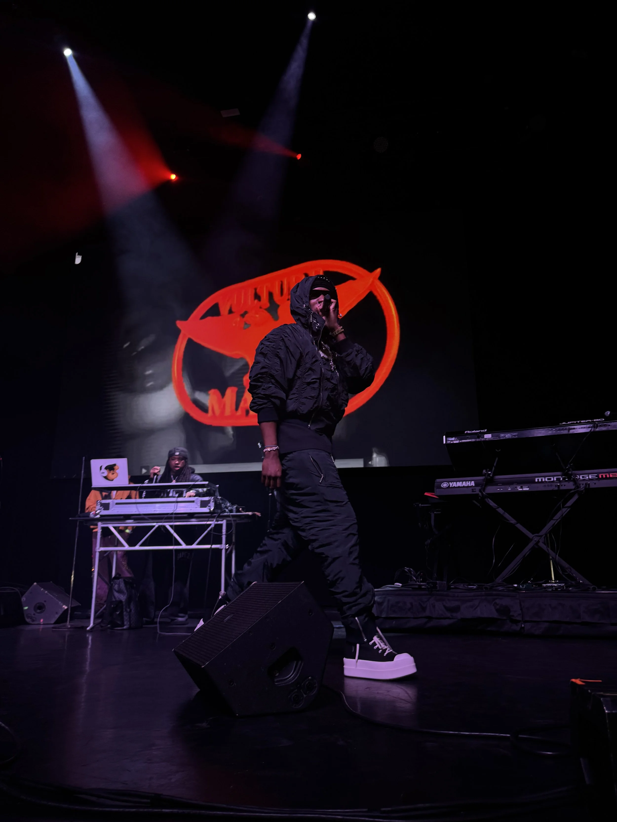A performer on stage wearing a hooded outfit and performing in front of a DJ setup. The background features a red and orange graphic with spotlights illuminating the scene.