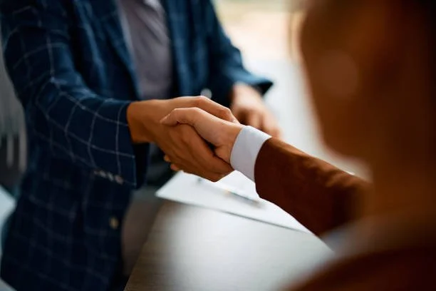Close-up of two people shaking hands in a business setting, with one wearing a plaid jacket and the other in a brown suit.