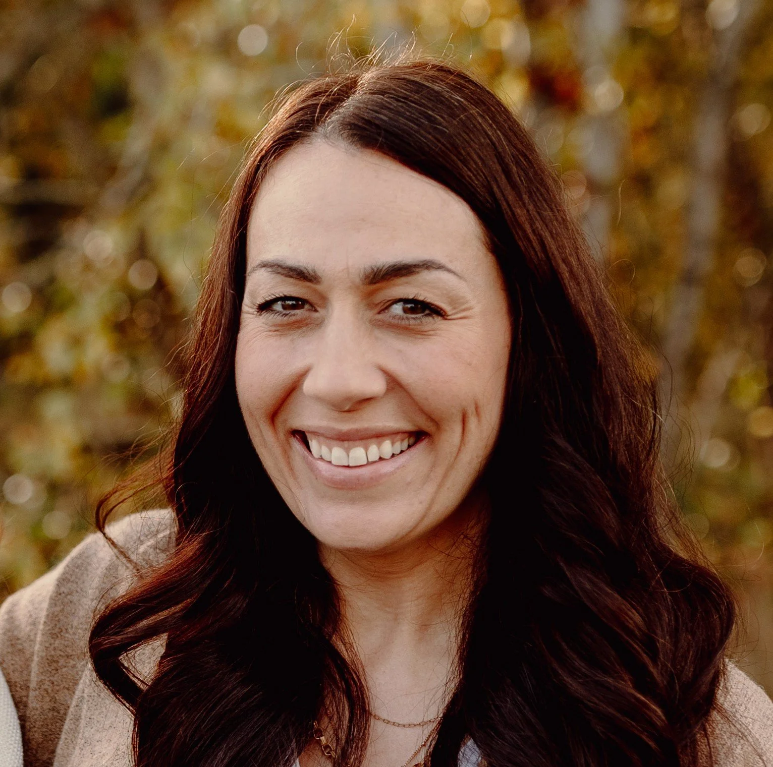 A woman with long dark hair smiling outdoors with a background of fall foliage.