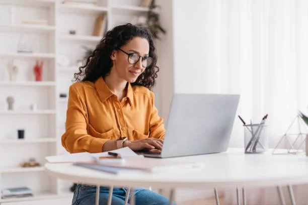 A woman wearing glasses and a mustard-colored shirt is working on a laptop at a white table, in a well-lit room with shelves in the background.