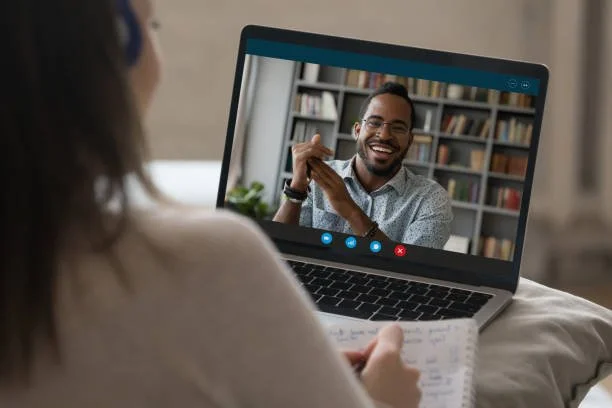 Person having a video call on a laptop, with a smiling individual on the screen and a notebook in hand.
