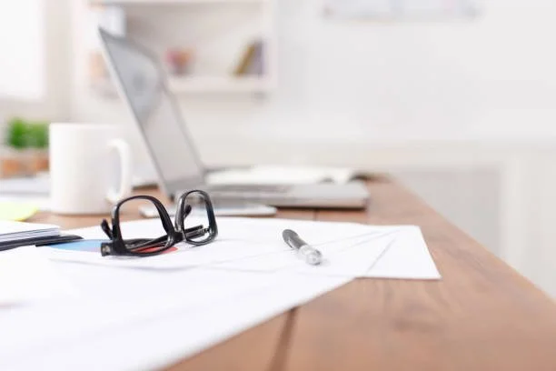 Office desk with glasses, pen, papers, and a laptop in the background.