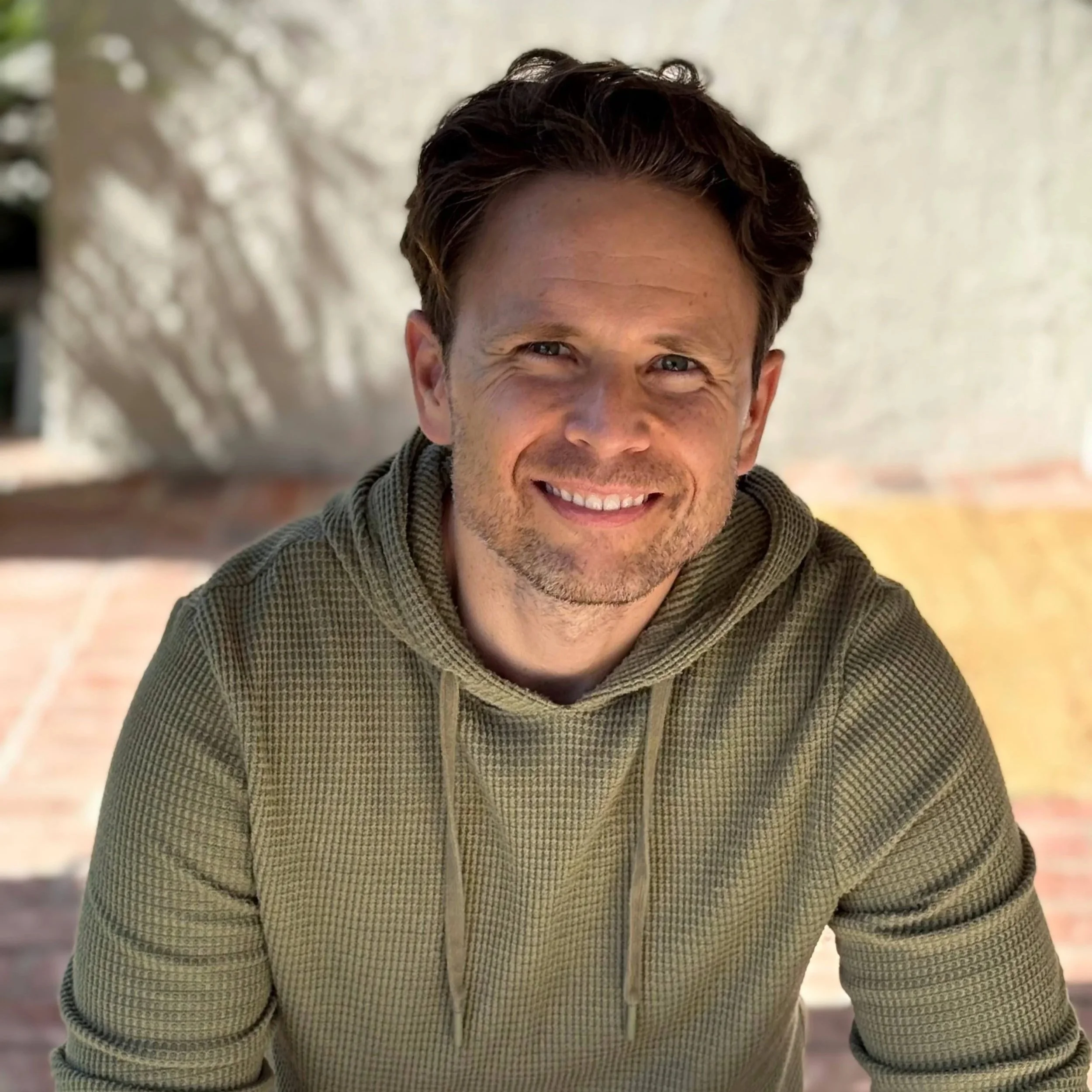 A smiling man with short brown hair and a light beard, wearing a green hoodie with drawstrings, outdoors with a blurred background of a wall and trees.