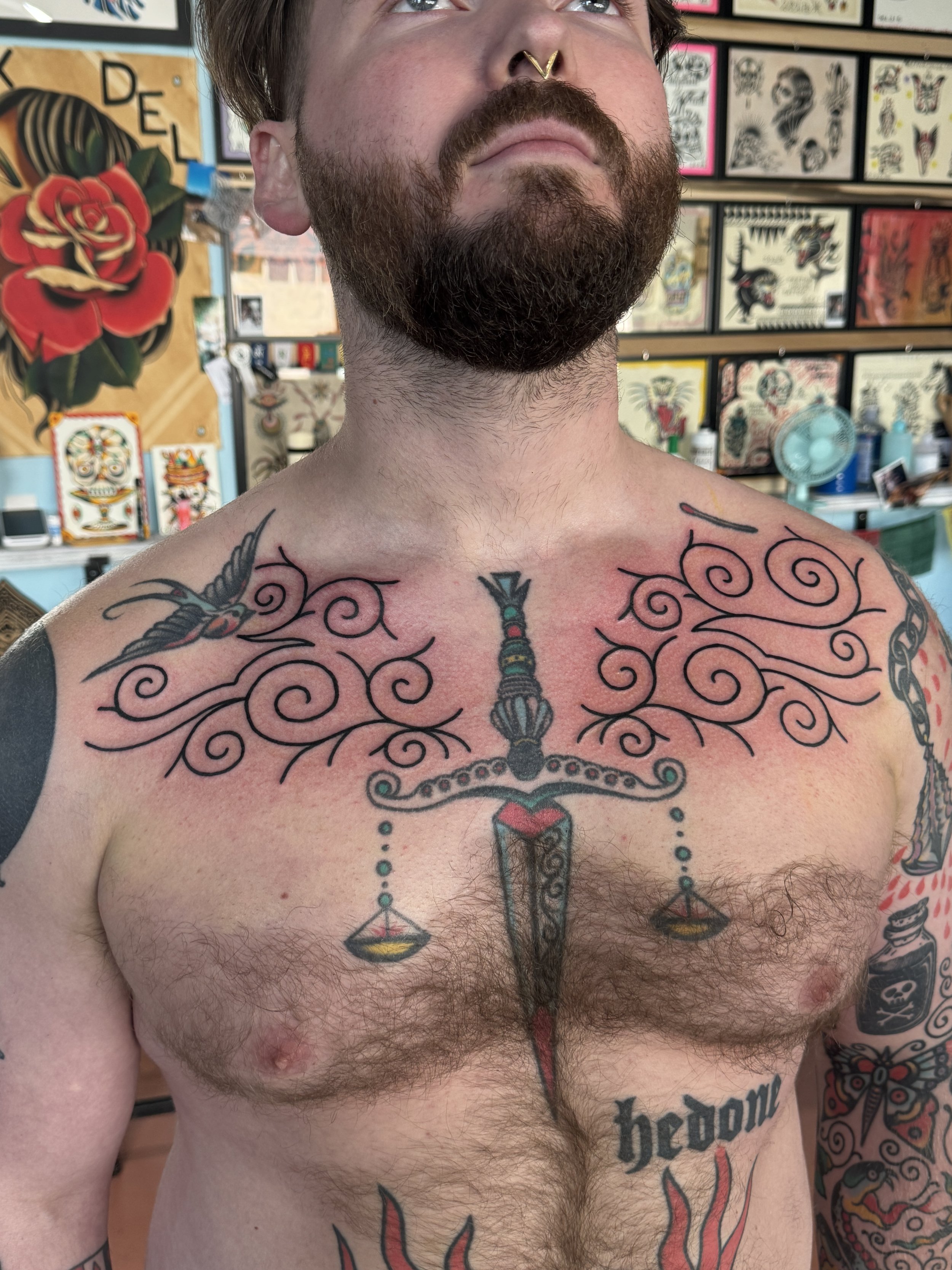 Man with a beard and multiple tattoos, including a large ornate sword with scales hanging from it and the word "hedone" on his chest, in an tattoo shop with artwork on the walls.