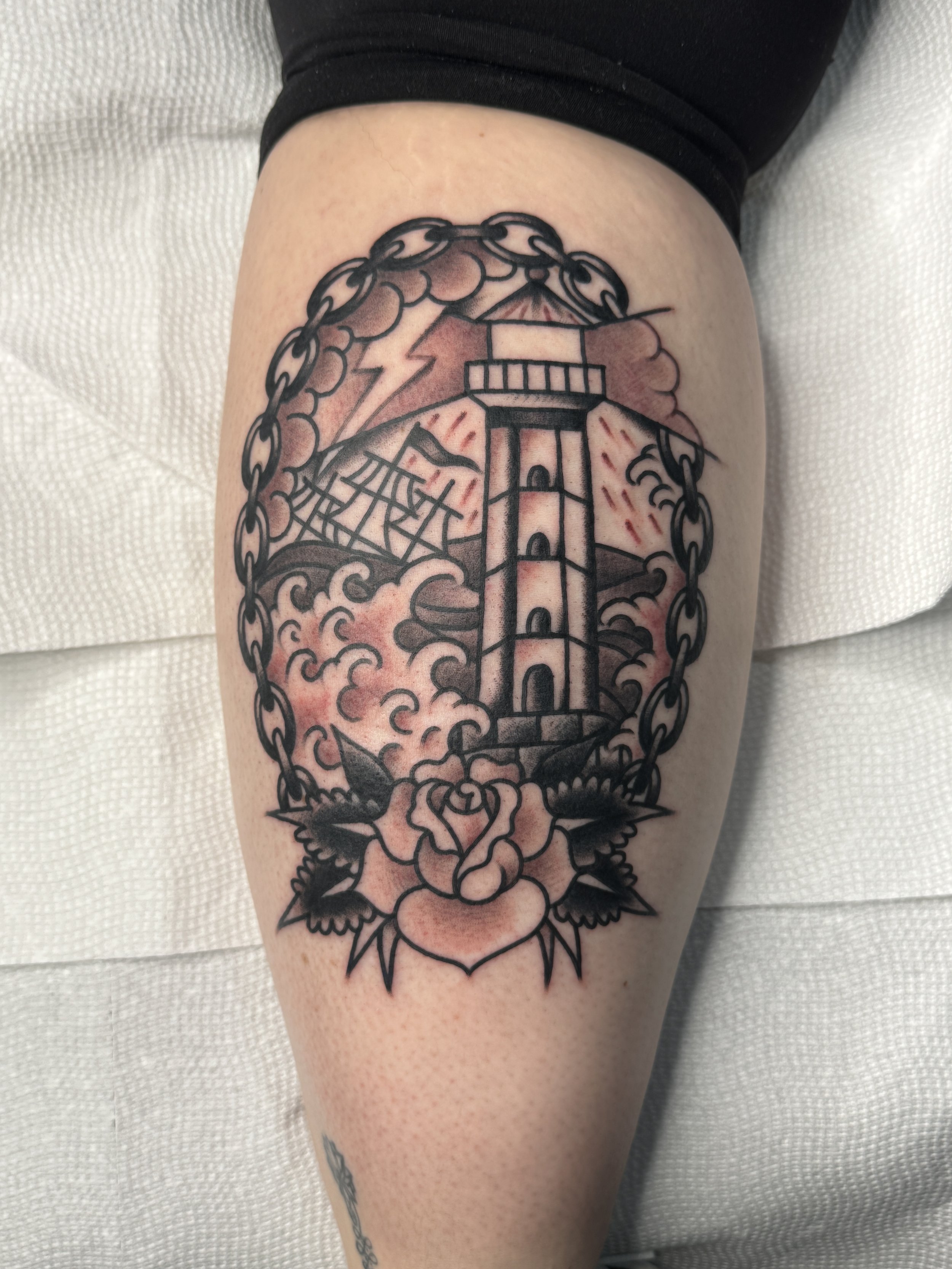 Tattoo of a lighthouse surrounded by clouds, ships, a chain, and roses on a person's thigh.