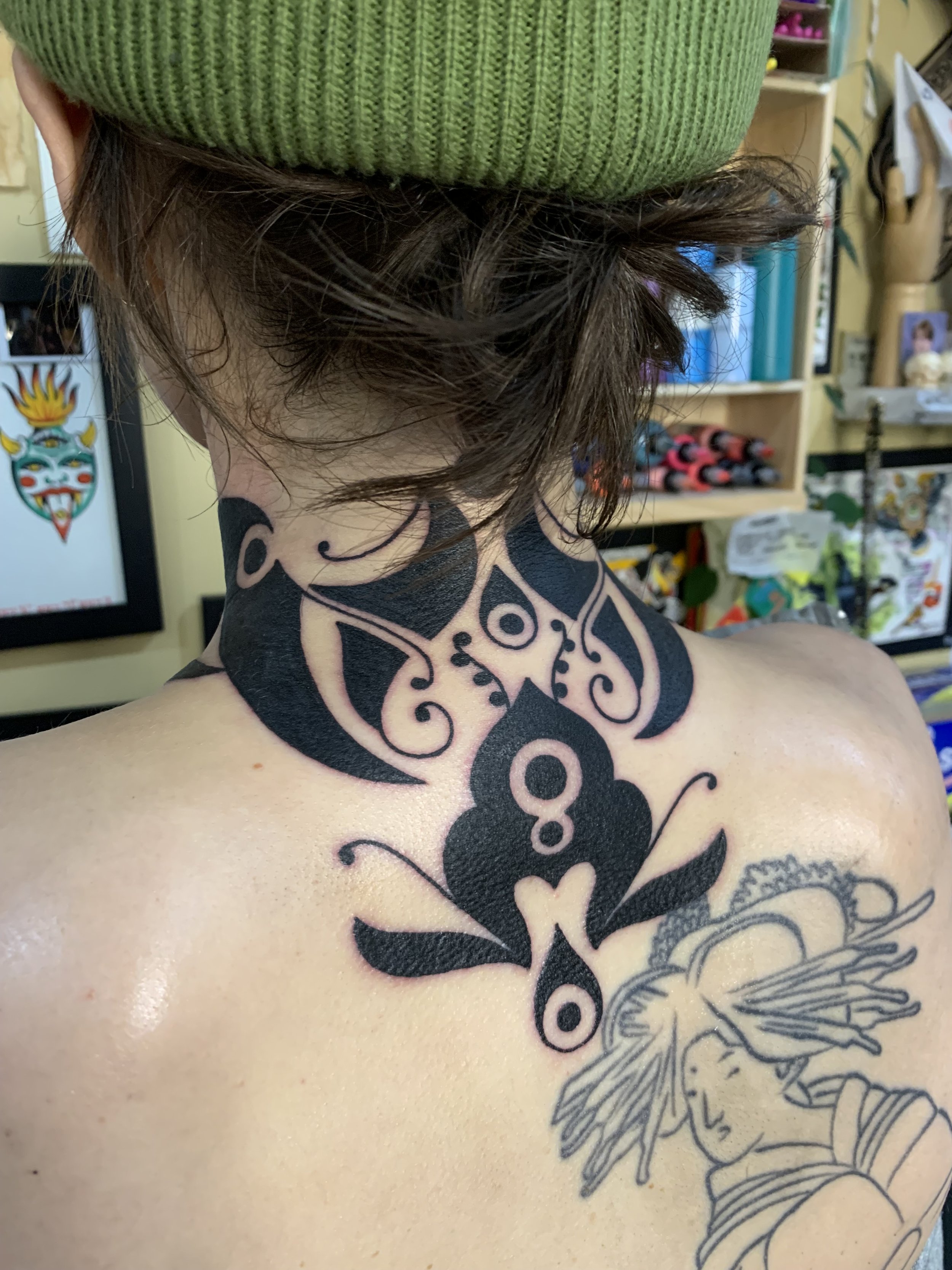 A person with an elaborate black tattoo on their upper back and neck, wearing a green knit hat, with a colorful background showing various supplies and artwork.