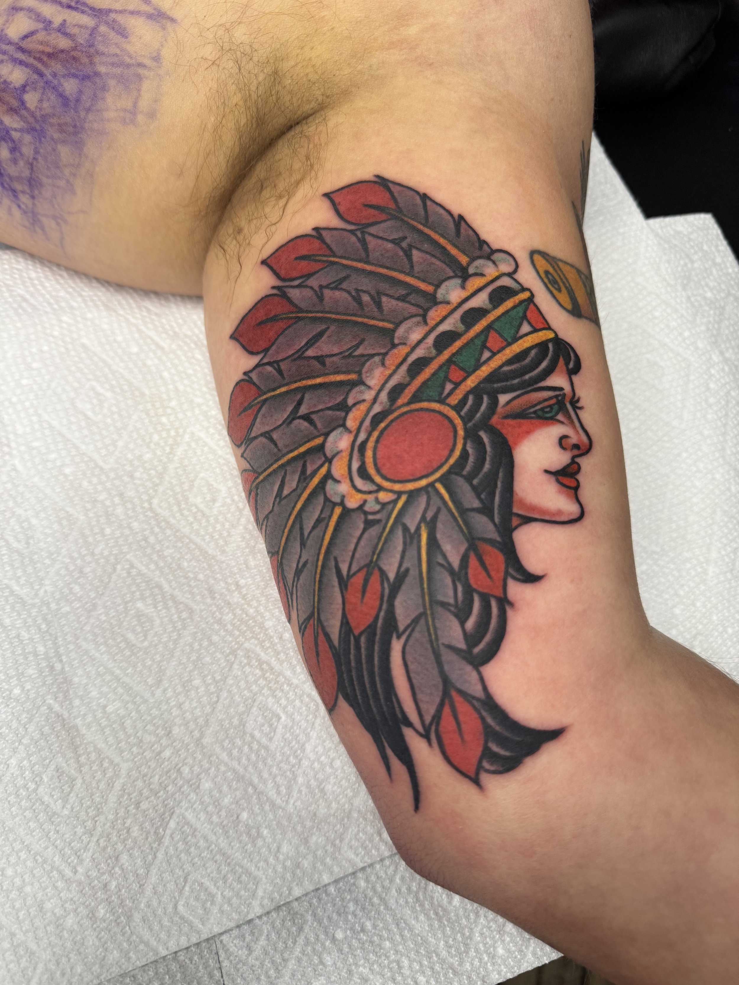 Tattoo of a Native American woman wearing a headdress with red, black, and gray feathers.