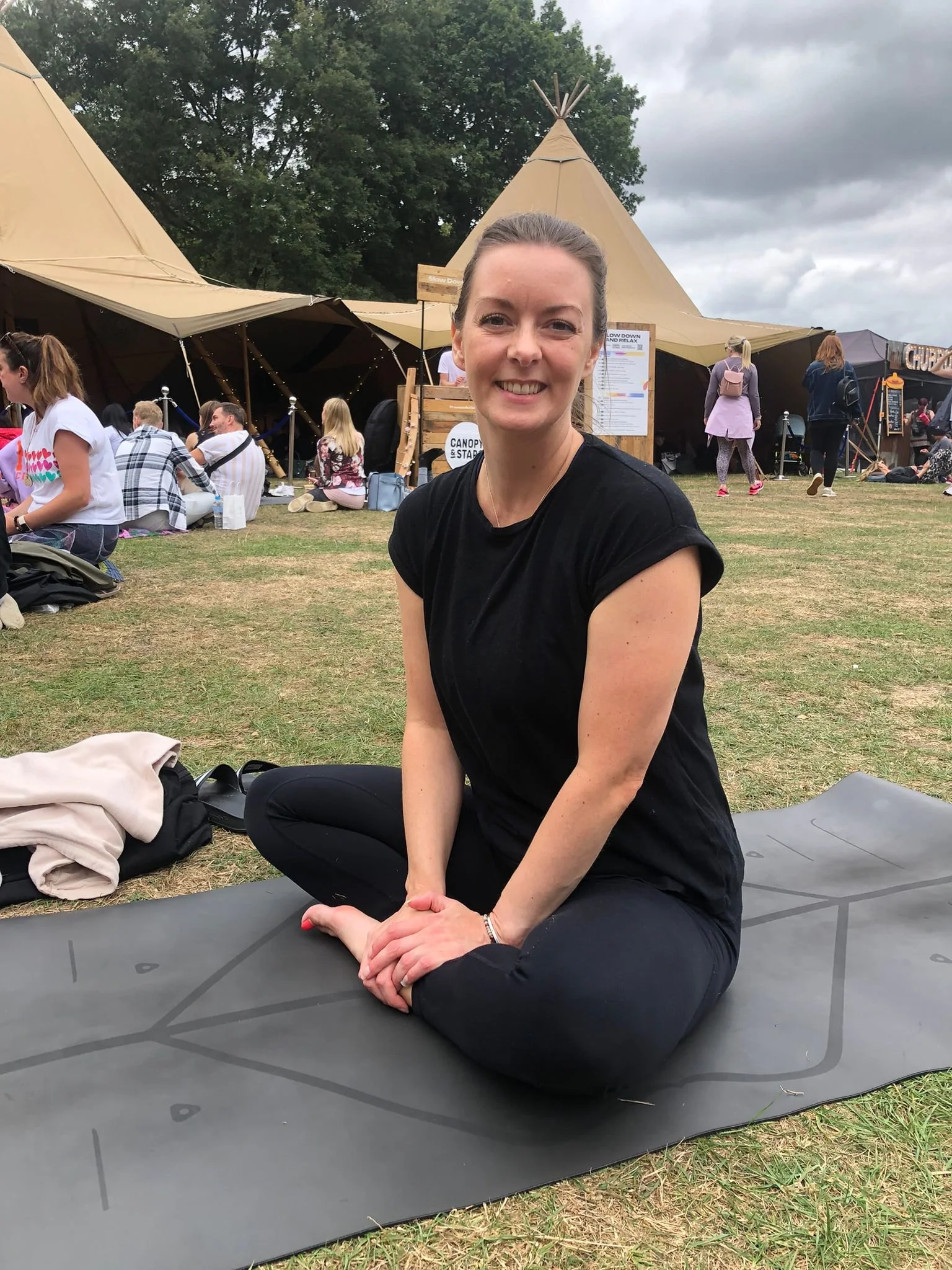 Stacey smiling and sitting cross-legged on a yoga mat outdoors, with large teepees and other people in the background, during a cloudy day.