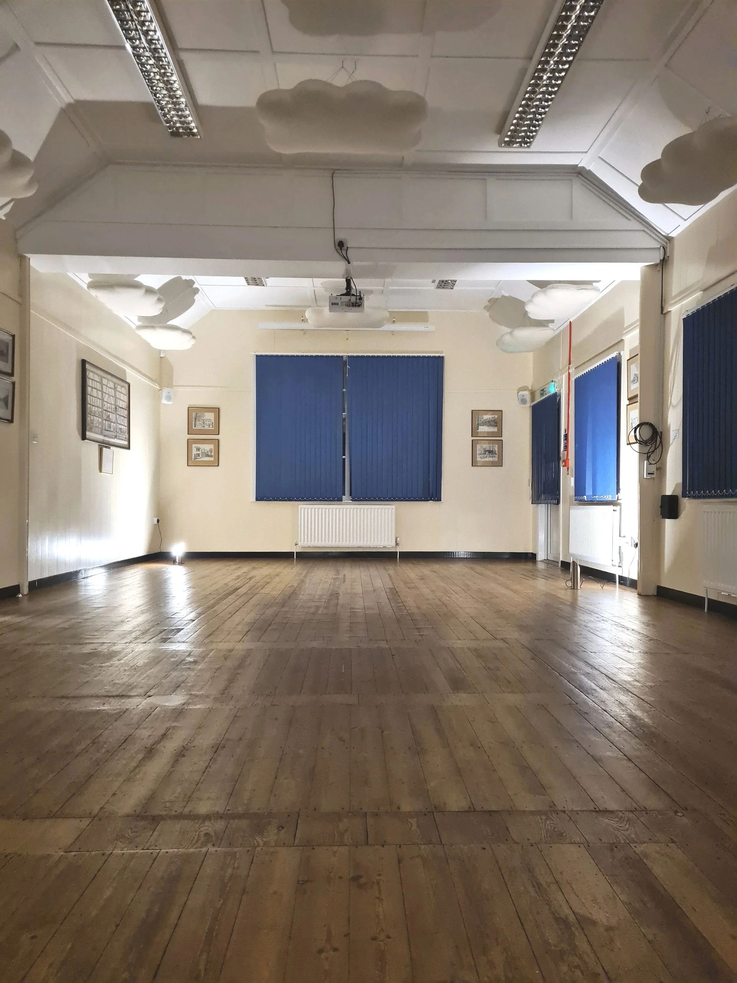 Empty room with wooden floor, framed pictures on the walls, large windows with blue blinds, a radiator, and a projector hanging from the ceiling.