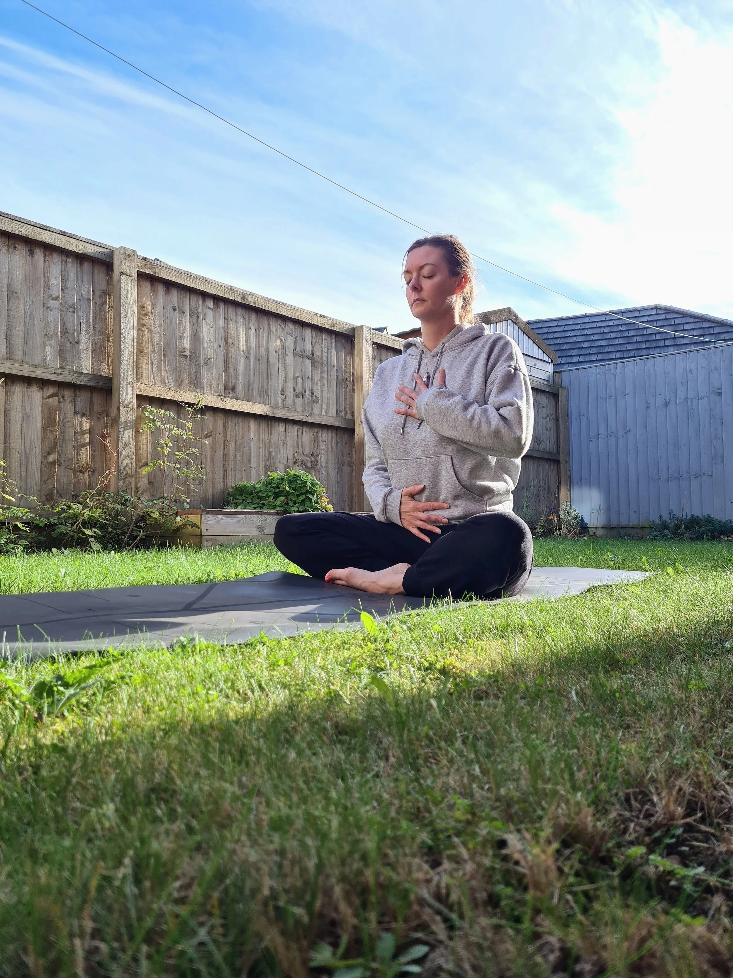 Stacey sitting cross-legged on a yoga mat in a sunny garden, practising mindful breathing with one hand on her chest and one on her abdomen.
